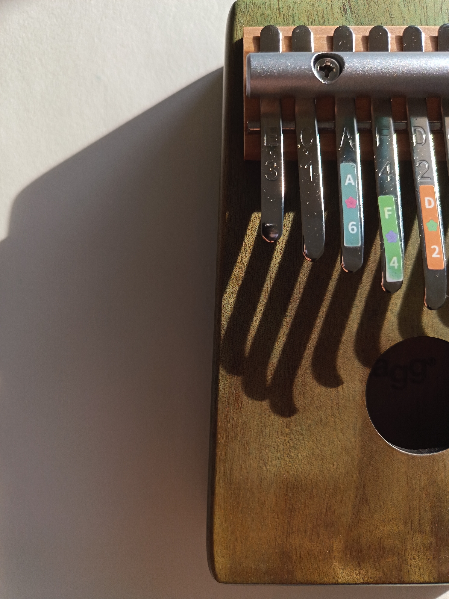 This is a close-up, high-angle, eye-level shot of a Kalimba, a thumb piano, resting on a light-colored surface. The Kalimba is oriented vertically, with its metallic tines facing the viewer and casting distinct shadows across the surface. The wooden body of the Kalimba has a greenish hue and a visible wood grain. Each of the tines is engraved with a letter and a number, and some also feature colored stickers with musical notes and digits. The background is out of focus, with a harsh shadow falling from the left side of the frame. The image does not depict any people or activities related to the city of Périgueux, France.