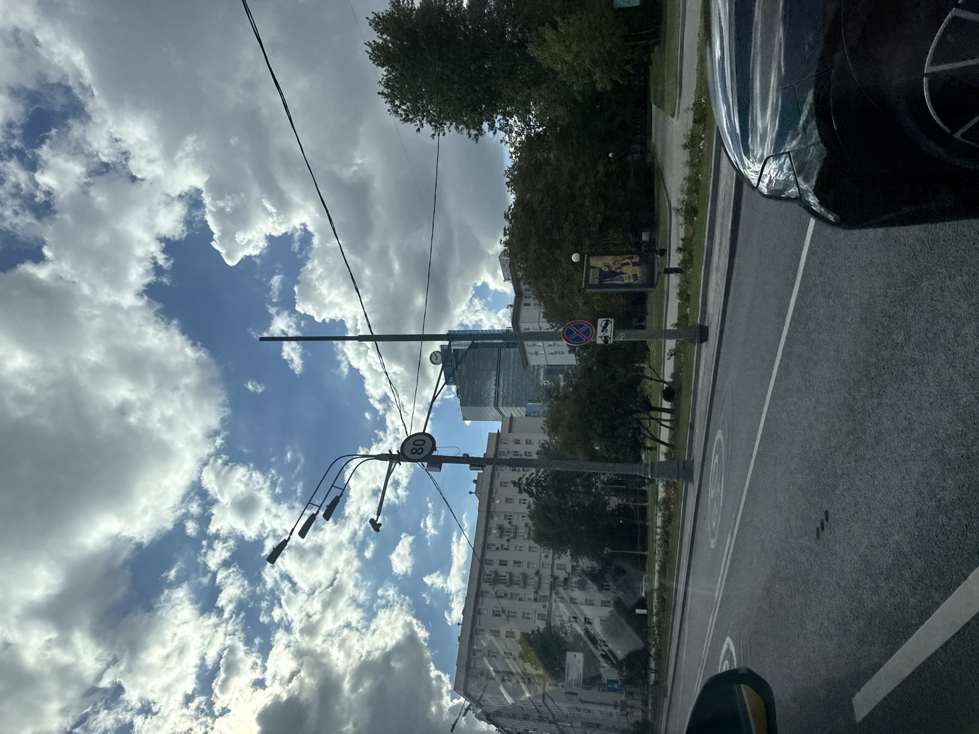 The image shows a street scene in Moscow, Russia, as viewed from inside a vehicle. In the foreground, the hood and fender of a dark-colored car are visible on the right, reflecting the sky. The road has lane markings and circular markings on its surface. A street lamp with multiple lights and a speed limit sign showing "80" is positioned to the left of the road. Behind the street lamp, a large, light-colored apartment building dominates the mid-ground, with many windows and balconies. Further in the background, a modern glass skyscraper with a distinctive shape stands out against the sky. Trees and foliage are visible along the side of the road and between the buildings. The sky is filled with dramatic clouds against a blue background. A blue and red traffic sign depicting a tow truck and a car is also visible on a pole.