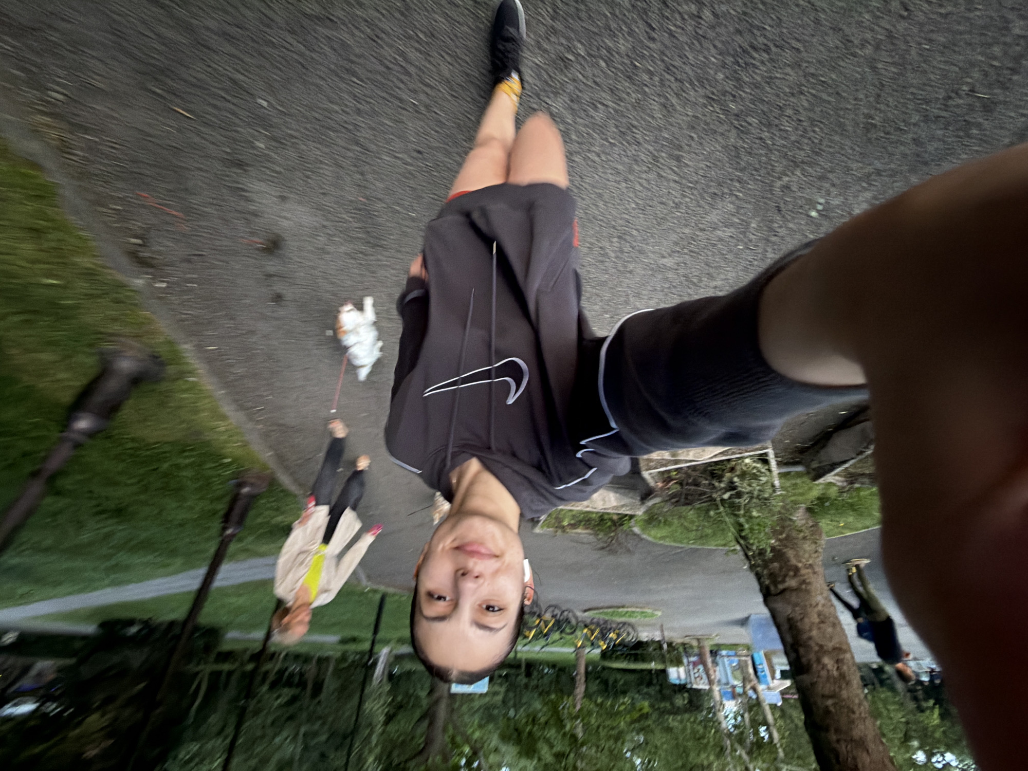 A young woman captures a selfie outdoors in what appears to be a park or paved pathway in Burgas, Bulgaria. The image is taken from a slightly high angle, looking down towards her body and the path ahead. She wears a dark gray hooded sweatshirt featuring a prominent white Nike swoosh logo on the front, dark shorts, black athletic shoes with yellow socks, and white wireless earbuds. Her expression is calm with a slight smile as she looks directly at the camera.
In the background, on the paved path, another person is seen walking a small, light-colored dog on a leash. This person wears a light-colored jacket over a bright yellow top and dark pants. Further back and to the right, partially obscured by a tree and the camera-holding hand, another individual in dark clothing is visible.
The setting includes areas of green grass, various trees, and paved asphalt paths. A rack of yellow rental bicycles is parked in the mid-ground to the right, and dark lampposts are visible to the left. The lighting is soft and diffused, suggesting it might be an overcast day, early morning, or late afternoon, rather than bright midday sun.