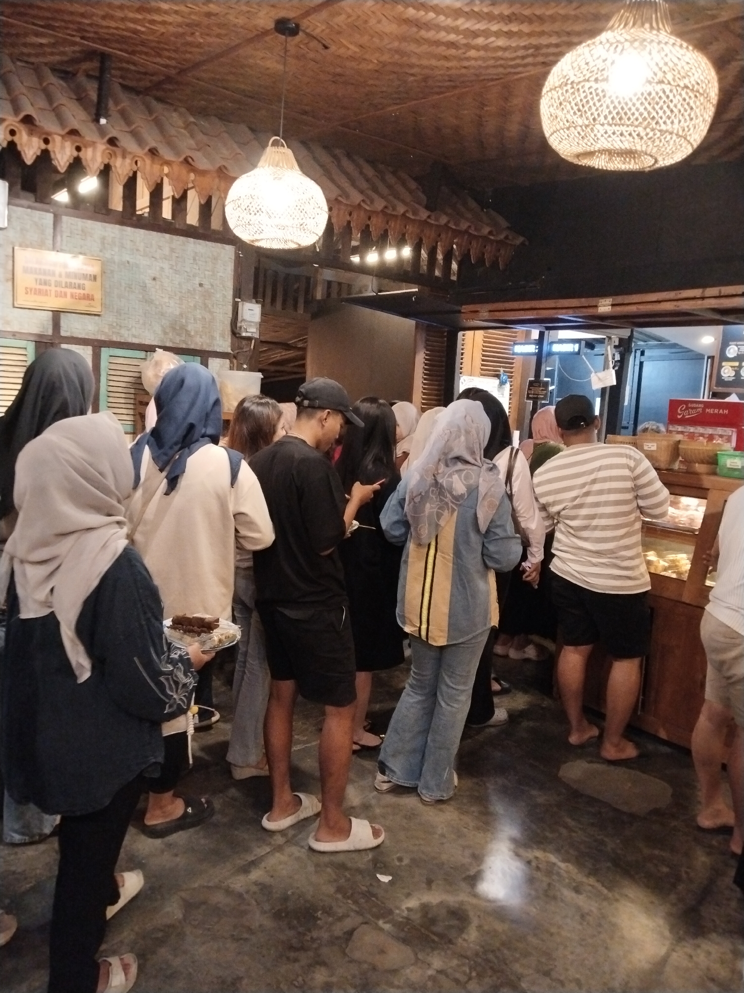 The image depicts a crowded indoor scene, likely a food establishment, with several individuals queuing. The overall ambiance suggests a casual setting. The flooring is a dark, textured material, possibly concrete or stone, with some reflective patches suggesting moisture or a polished surface. The walls appear to be constructed from natural materials, with a thatched or woven ceiling overhead. Decorative lamps, made from similar woven material, hang from the ceiling, providing warm, ambient lighting.
In the foreground, on the left, a person wearing a dark headscarf and black pants is holding a clear plastic plate filled with small, rectangular brown pastries. The person is wearing a dark long-sleeved top. To their right, another individual in a light blue headscarf and a light-colored long-sleeved shirt is partially visible. Further back, a person wearing a dark baseball cap, black t-shirt, and shorts is looking down, possibly at their phone or something on the floor. A person in a light blue and yellow striped denim jacket and blue jeans is standing in the middle ground, facing away from the camera. To the right, a person in a striped t-shirt and dark shorts is facing a display case filled with food items. The display case has a wooden frame and glass front, showcasing various baked goods. Behind this person, another individual in a white and light-colored striped shirt is also visible.
A sign with Indonesian text is mounted on the wall behind the individuals in the foreground. The text on the sign is partially obscured but appears to be a set of regulations or rules. In the background, there is a counter with more food displays and signage, including a red sign with "GUDANG SARAM MERAH" written on it. The overall scene suggests a busy environment where customers are waiting to be served. The presence of people dressed in casual attire and the nature of the establishment imply a public gathering place, possibly a bakery or cafe in Kecamatan Pati, Indonesia.