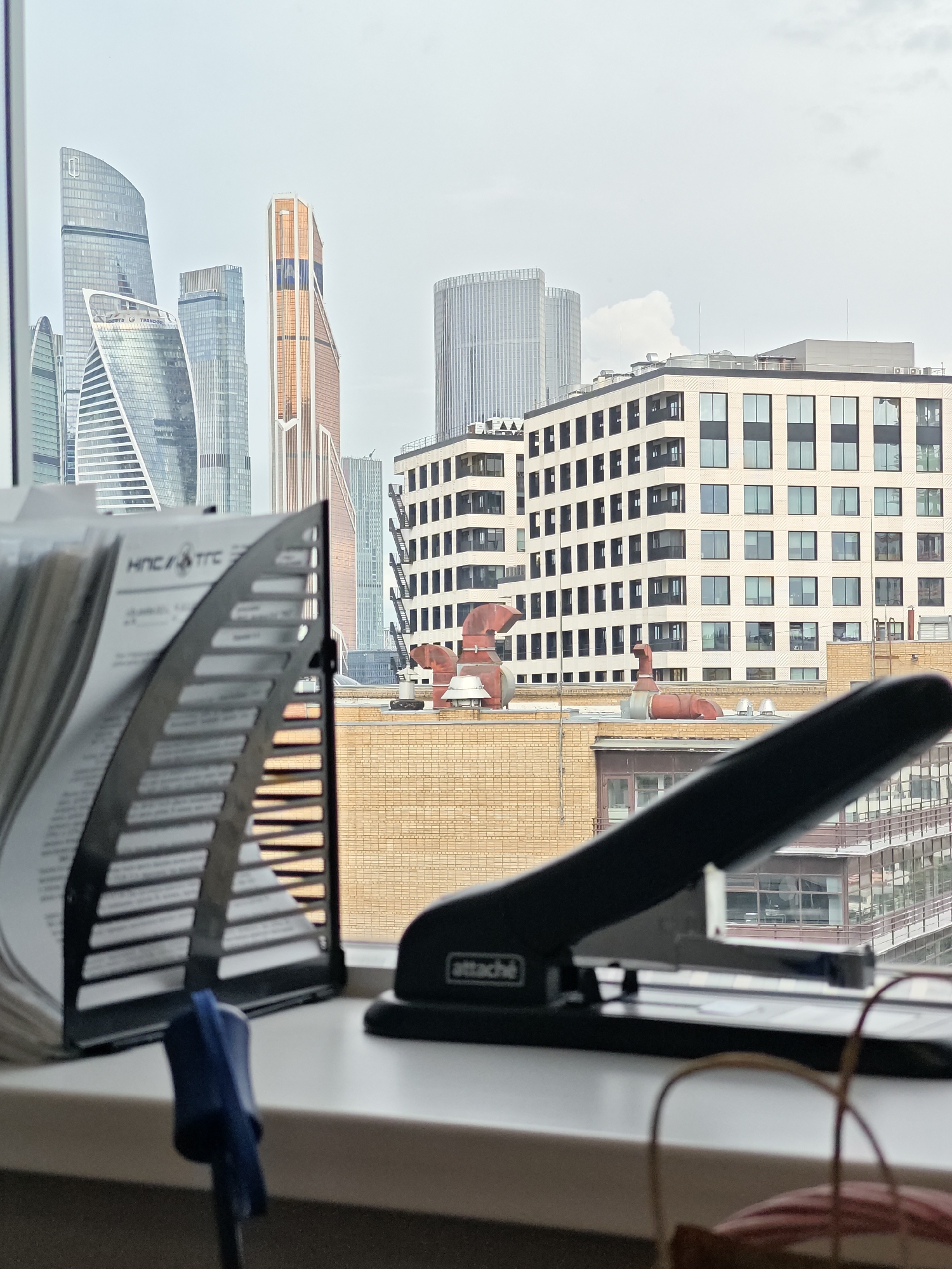 The image depicts a view of the Moscow International Business Center, also known as Moscow-City, from an indoor vantage point, likely an office. In the foreground, a black stapler and a document holder are placed on a white surface, possibly a windowsill. The stapler has the brand name "attaché" visible. The document holder contains papers with Cyrillic text. Beyond the foreground objects, a cityscape unfolds, dominated by numerous modern skyscrapers. Notable buildings include the Federation Tower, Neva Towers, and Triumph Palace, all characteristic of Moscow-City's contemporary architecture. The sky is overcast, suggesting a cloudy day. The scene captures an urban environment with a blend of office supplies and a distant panoramic view of a major business district.