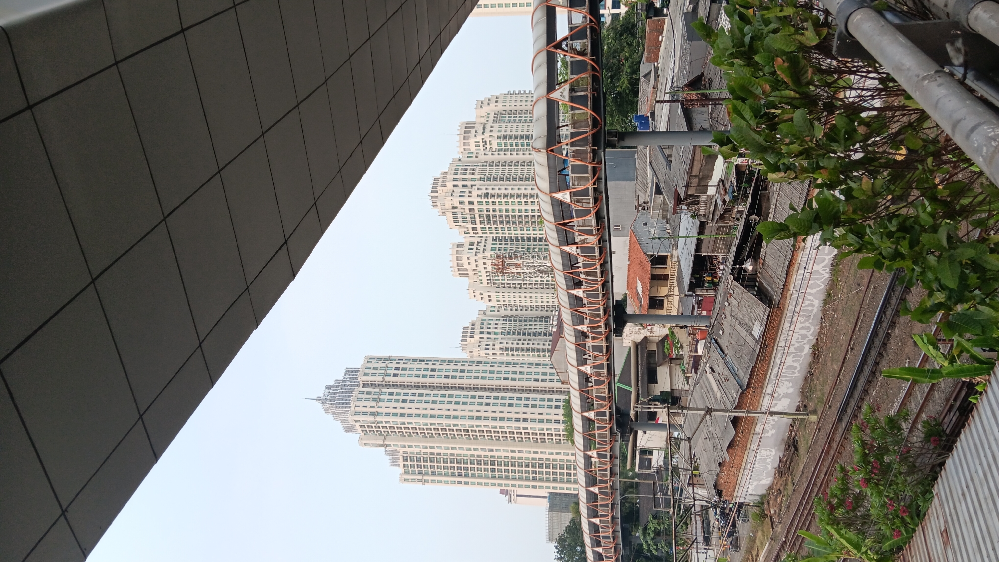 The image depicts a city scene in South Jakarta, Indonesia, dominated by modern high-rise buildings and an elevated pedestrian walkway. In the foreground, a grey, textured ceiling or awning extends across the top, partially obscuring the sky and the view of the buildings. Below this, a network of train tracks runs horizontally across the middle ground, flanked by low-lying buildings with corrugated iron roofs and some greenery.
The most prominent structures are a cluster of tall, white apartment buildings in the background, featuring numerous windows and a distinct architectural style with tiered tops. Between the train tracks and the apartment buildings is an orange and grey pedestrian bridge, supported by pillars, which spans the width of the frame. The area beneath the bridge appears to be a mix of residential and commercial structures. The overall atmosphere is urban, with a blend of old and new infrastructure. There are no visible people in the image.