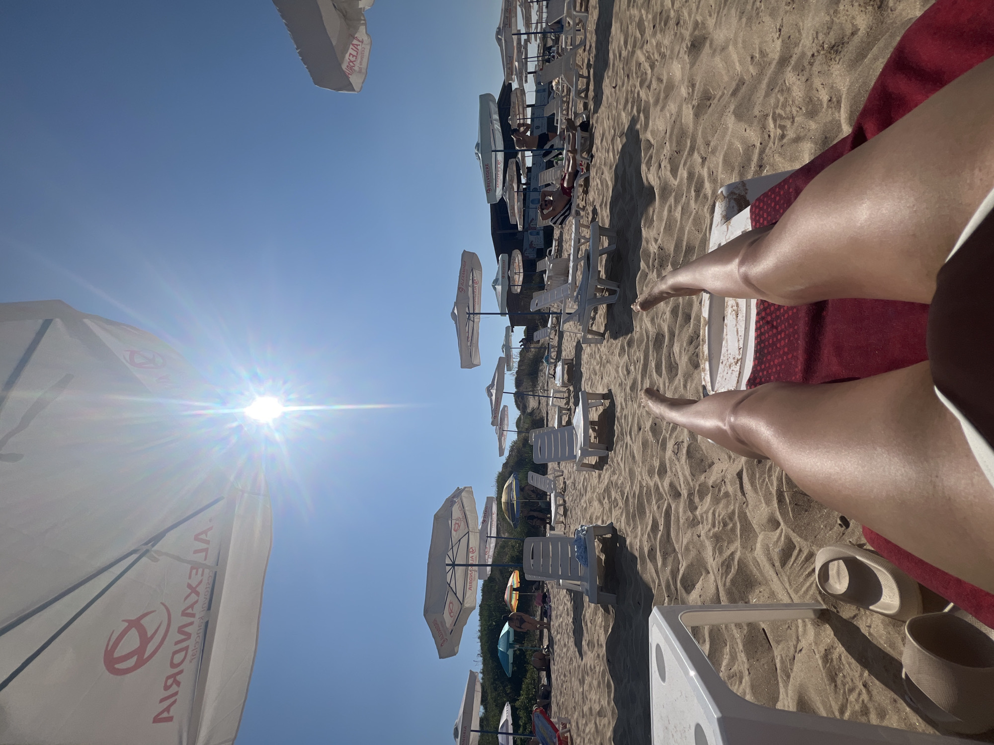 The image shows a sun-drenched beach scene in Primorsko, Bulgaria. In the foreground, a person's tanned legs are stretched out on a red towel covering a white lounge chair. A white side table is to the left, with a pair of beige slides on the sand nearby.
The beach is covered in fine sand, with numerous white lounge chairs and umbrellas dotting the landscape. The umbrellas display the logo "ALEXANDRIA" and "BULEVARD". In the background, a low building with a dark roof is visible, likely a beach bar or restaurant. Several people are scattered across the beach, some sitting on chairs, others standing or walking. The sky is a clear, bright blue, with the sun intensely shining, creating lens flare and casting sharp shadows. The overall atmosphere is one of a relaxed and sunny day at the seaside.