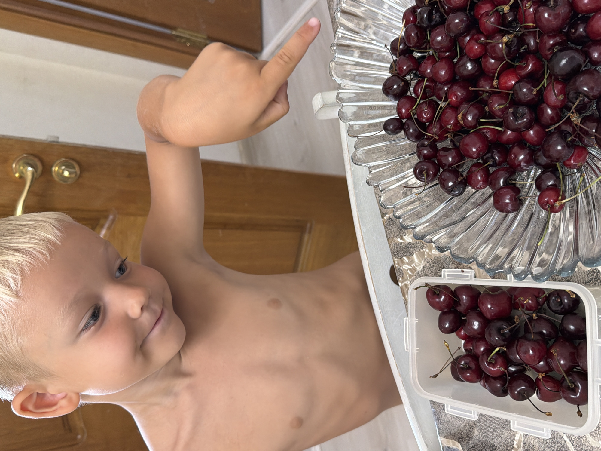 A young boy with blond hair and blue eyes, wearing no shirt, is leaning over a table with two containers of cherries. He is pointing to the cherries in a clear glass bowl with his right index finger. The bowl is filled with dark red cherries. To his left is a white rectangular plastic container also filled with cherries. The boy has a slight smile and his head is tilted down, looking at the cherries. The background shows two wooden doors, one with a brass doorknob and the other ajar. The scene appears to be indoors, possibly in a home. There is no indication of the city of Sochi, Russia, in the image.