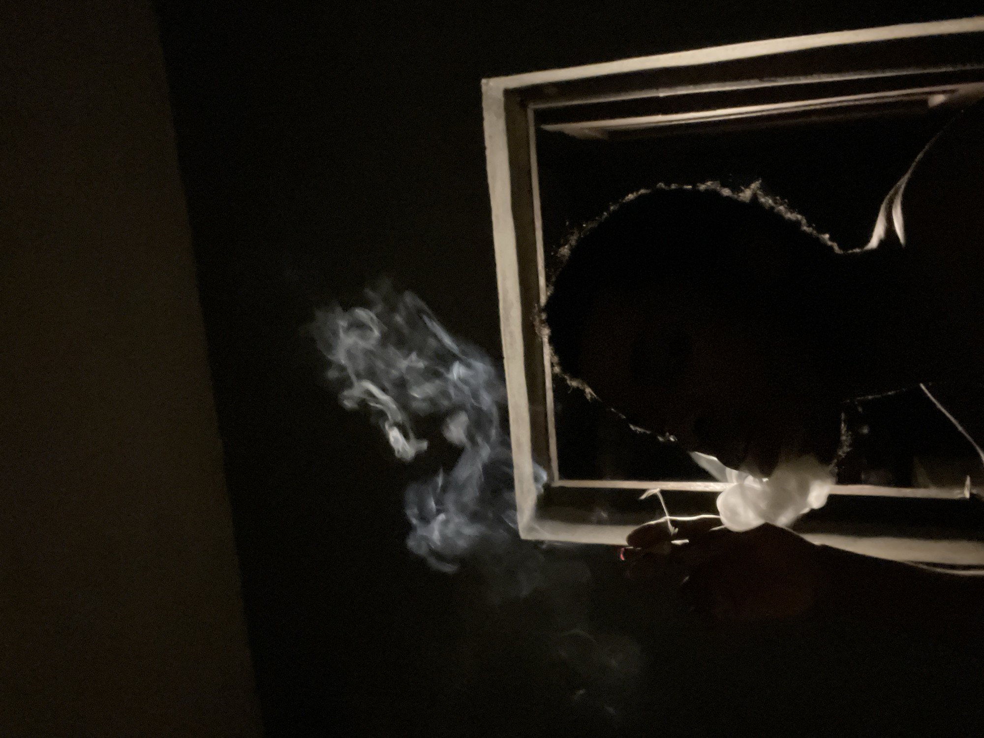 A silhouetted person is smoking indoors, with smoke billowing towards the left of the frame. The person is holding a lit cigarette. The individual is framed by a window. The location is Nkpor, Nigeria. The scene is very dark.
