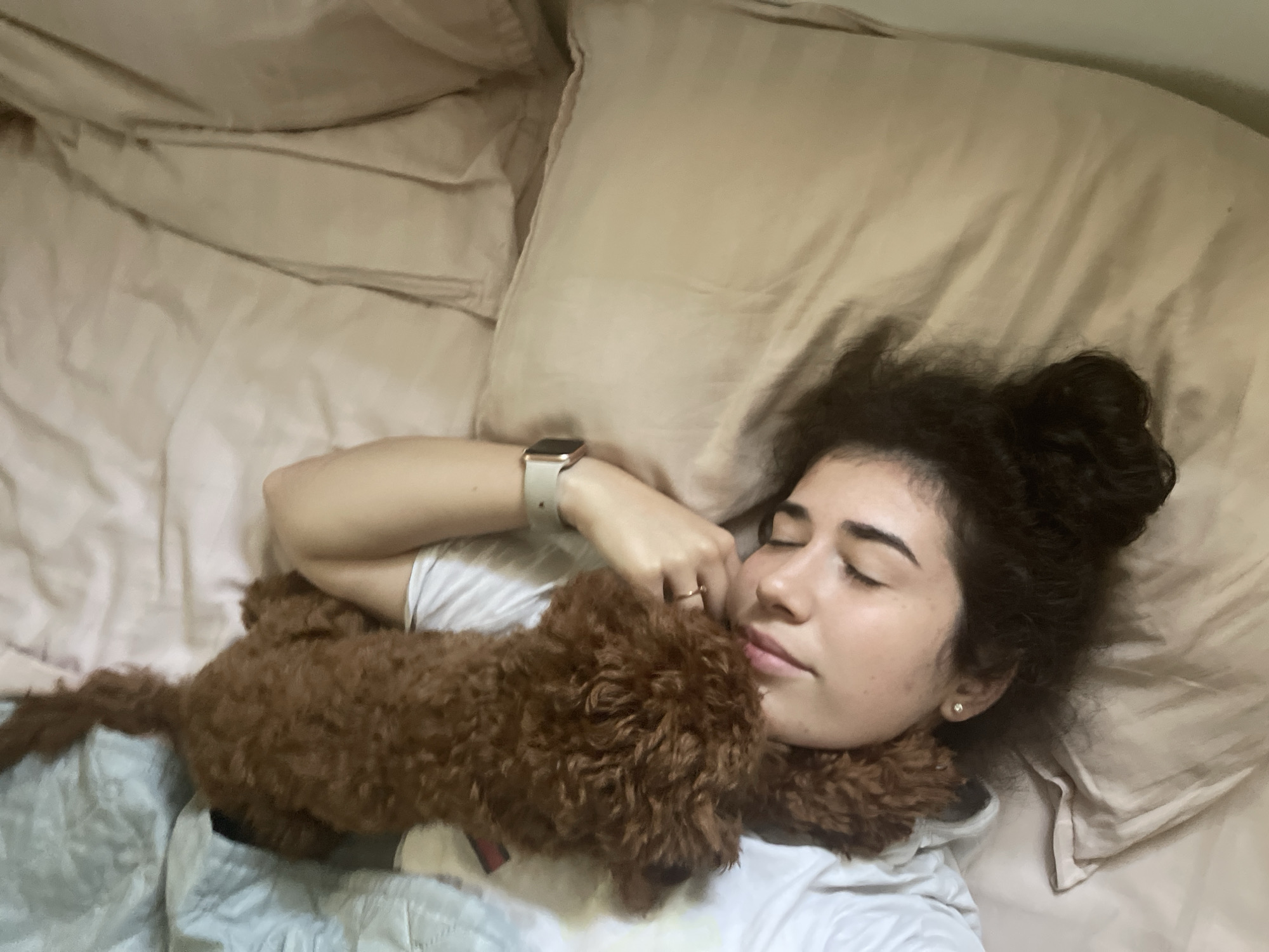 The image depicts a close-up aerial view of a young woman lying in a bed, with a small, brown, curly-haired dog on top of her, nuzzling her face. The woman has her eyes closed and her head is turned to the side, resting on a pillow. She is wearing a white top and a smartwatch on her left wrist. The dog appears to be a poodle or poodle mix. The bedding is a light beige color and appears to be made of a silky material. The background is blurred, suggesting a shallow depth of field. There is no indication of the location being in Sofia, Bulgaria.
