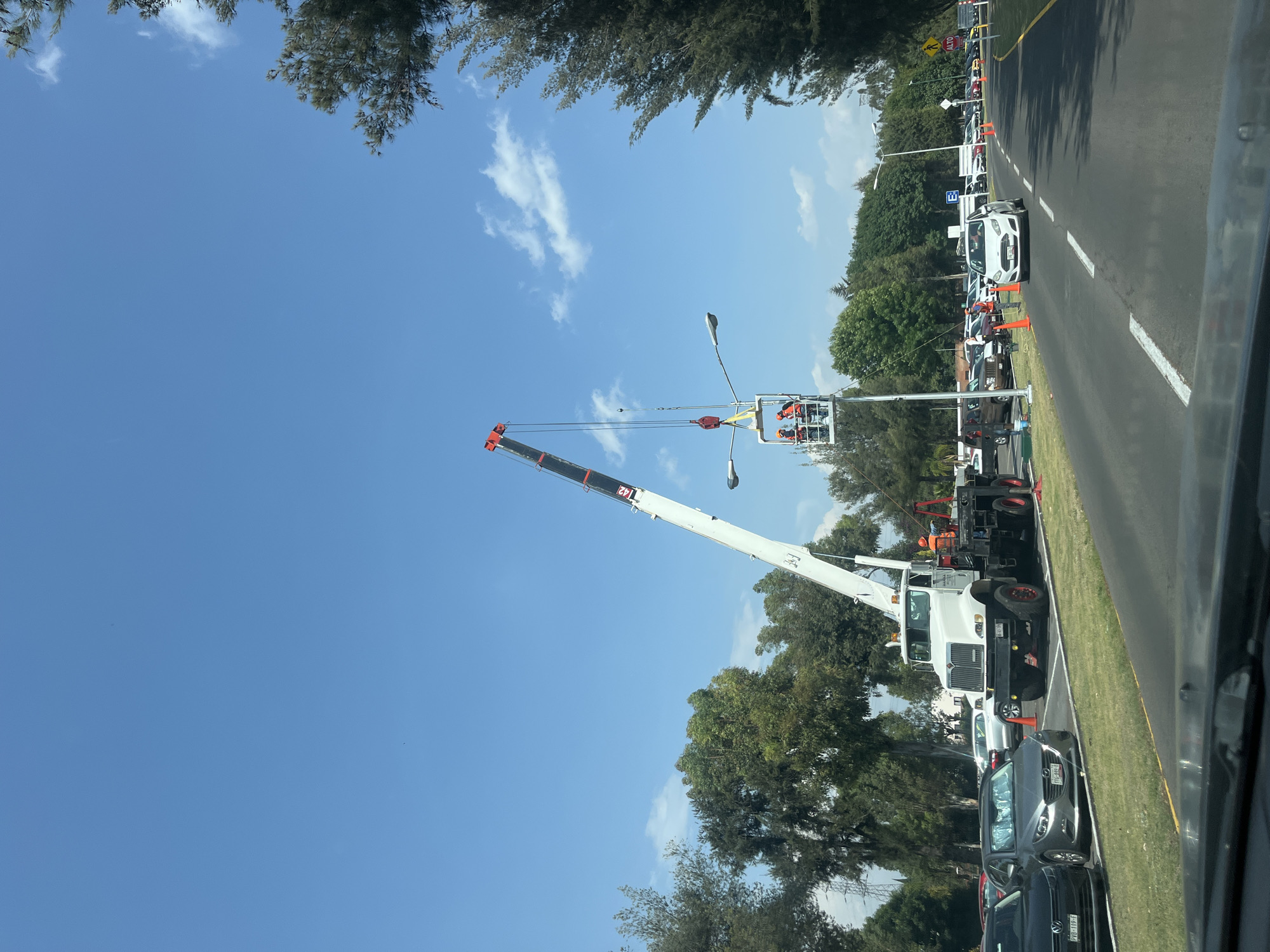 The image depicts a street scene in San Andrés Cholula, Mexico. A large white crane truck is parked on the side of the road, its boom extended upward, holding a personnel lift containing two workers near a street light. Multiple cars are parked along the side of the road behind the crane, and orange traffic cones are placed along the line of vehicles. A road runs parallel, where other cars appear to be passing. Trees line the street, and a clear blue sky with some clouds forms the backdrop.