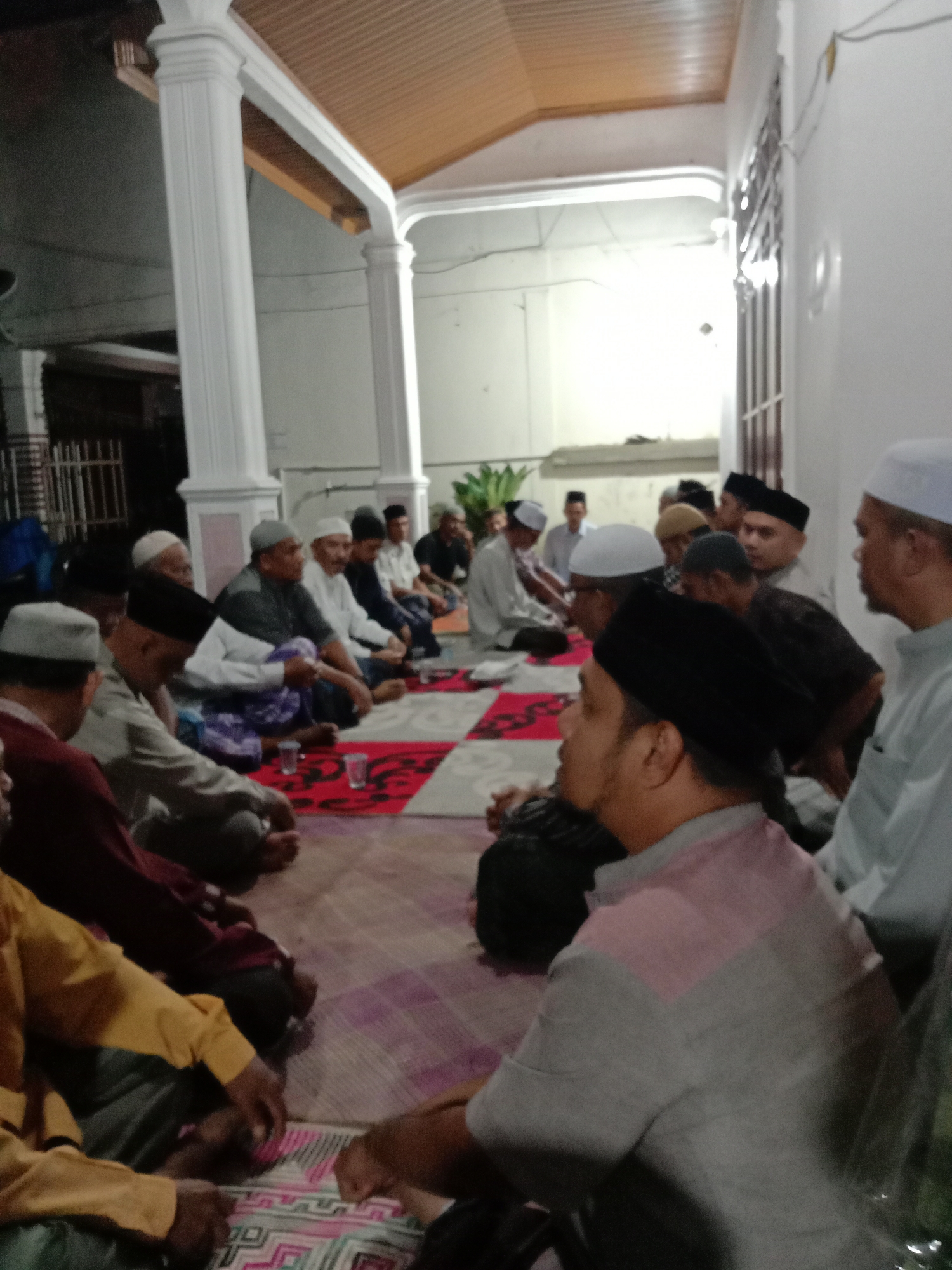 A group of men, presumed Muslim based on the headwear, are sitting on the floor of a building in Lhokseumawe, Indonesia. They are in an indoor-outdoor space, part of which is under a roof supported by white columns. They are on floor coverings of varying designs including one patterned in red and white and another in pink and purple. Some have glasses of water in front of them. The group appears to be engaged in a meeting or gathering. The building is painted white.