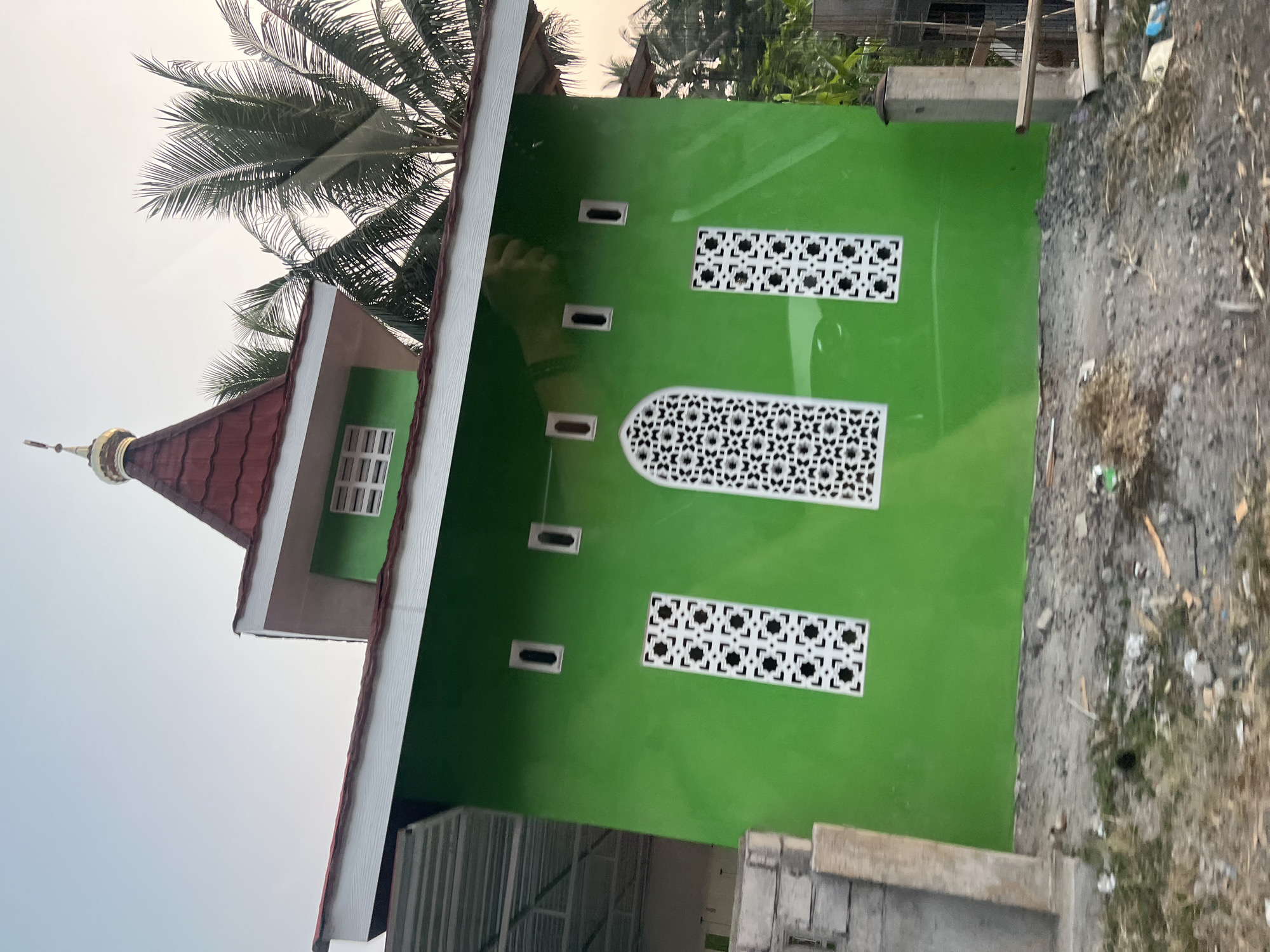 The image shows a small, bright green building with a red tiled roof and a golden dome, likely a mosque, in Kapulogo, Indonesia. The building has several decorative white windows with intricate geometric patterns. In the background, palm trees and other vegetation are visible, as well as a cloudy sky. The foreground shows a dirt and gravel area with some debris. A portion of a metal roof structure is visible on the left side of the building, suggesting it might be part of a larger complex or under construction. There are no people visible in the image.