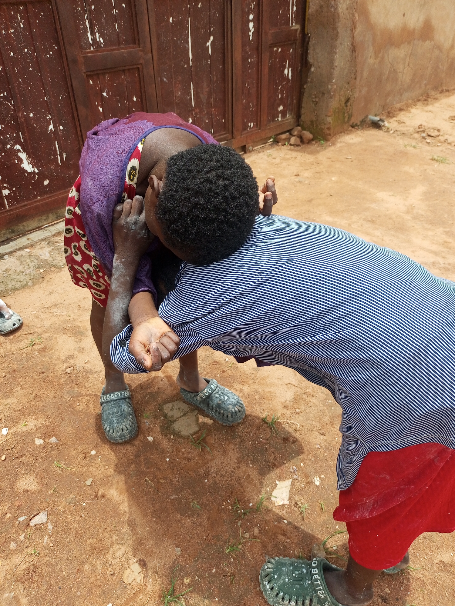 Two individuals are engaged in an interaction, with one person bending over and the other leaning towards them, their faces obscured from view. The person on the left is wearing a purple garment with a patterned hem and dark pants. The person on the right is wearing a blue and white striped shirt and a red skirt or wrap. Both individuals are wearing dark, open-toed sandals. The ground is covered in reddish-brown dirt with scattered stones and sparse green vegetation. In the background, there is a weathered wooden door with white paint splatters and a rough, tan-colored wall. The overall scene suggests an outdoor setting in what appears to be a village or rural area in Tempure, Nigeria. The lighting indicates it is daytime.