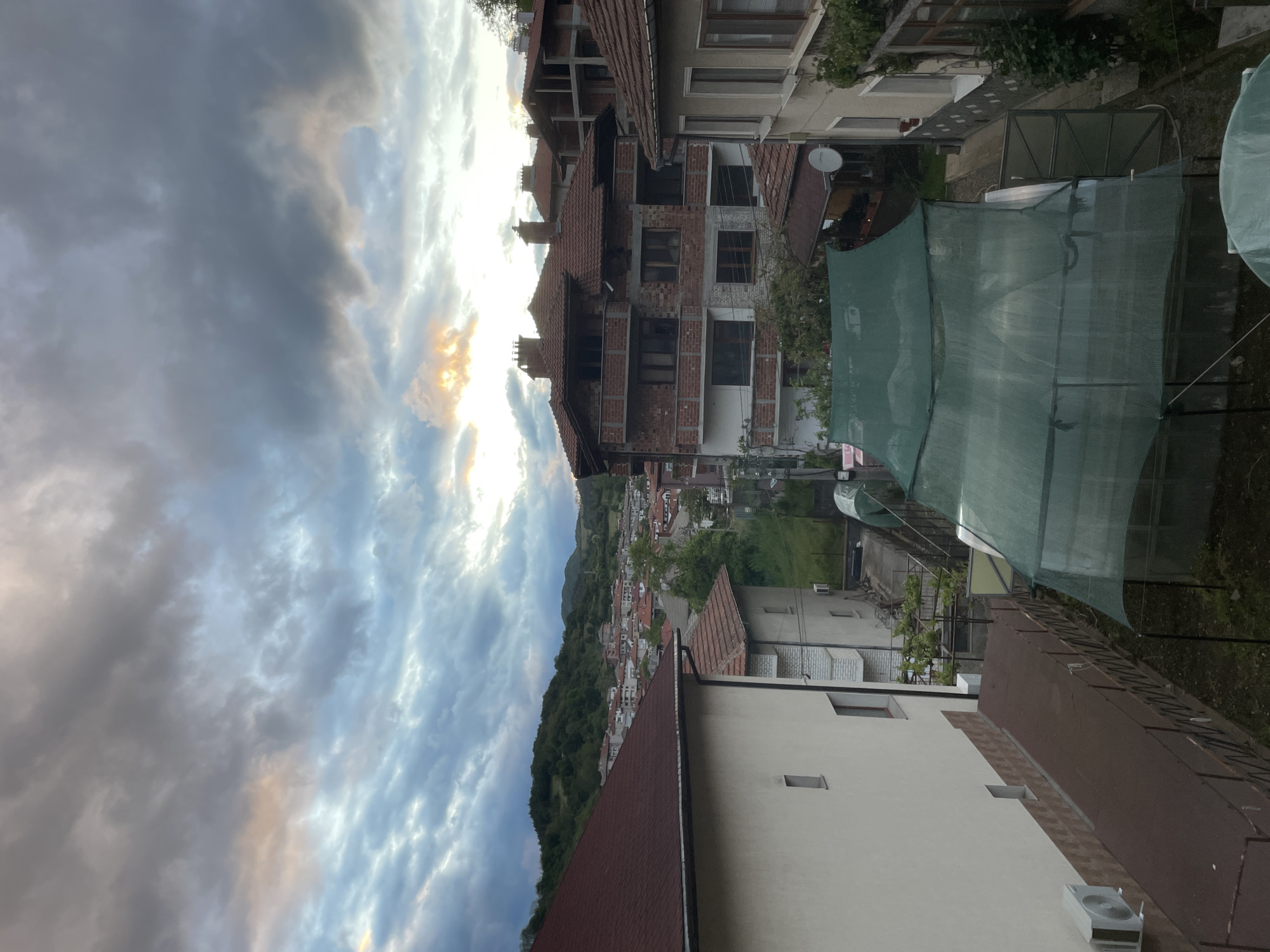 This is an exterior shot of the city of Zlatograd, Bulgaria, taken from an elevated position. The sky is filled with dramatic, swirling clouds, hinting at either sunrise or sunset. The buildings are of various construction types; some are brick, others are stucco or plaster, and they all have roofs of red tile. A large section of a structure covered with a green material dominates part of the frame. The city appears nestled in a valley, with forested hills in the background. An air conditioning unit is mounted on one of the buildings. There are no people visible.