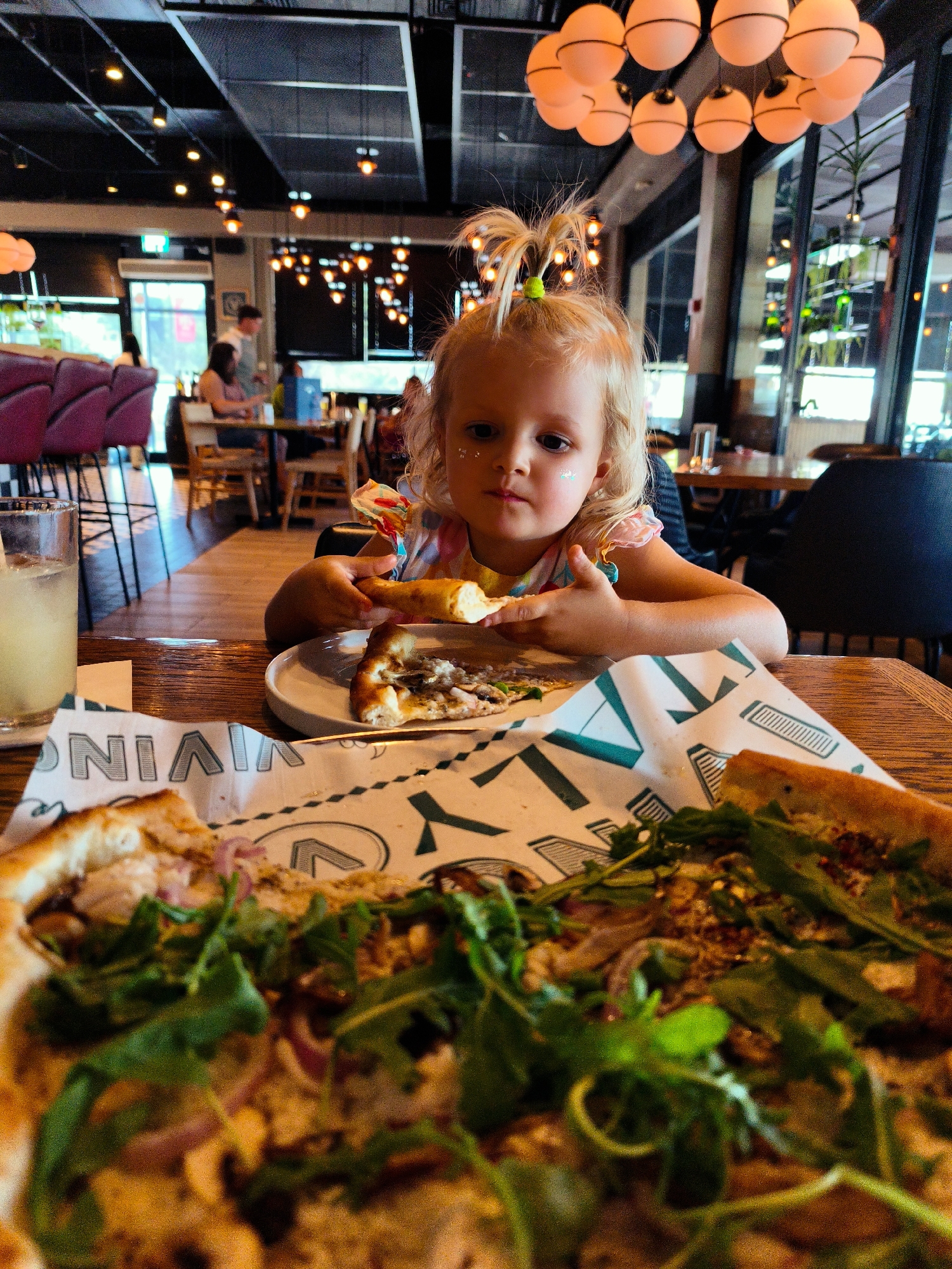 A young girl with blonde hair tied in a messy ponytail and glitter on her cheeks is sitting at a wooden table in a restaurant. She is holding a slice of pizza and looking directly at the camera. In the foreground, there is a large pizza with arugula and red onions, and a glass of a yellow beverage. The background shows a dimly lit restaurant interior with light fixtures hanging from the ceiling and other patrons seated at tables. The restaurant appears to be modern with a dark ceiling and large windows. The overall scene captures a casual dining experience with a focus on the child and the food. The location is a restaurant in Be'er Sheva, Israel.