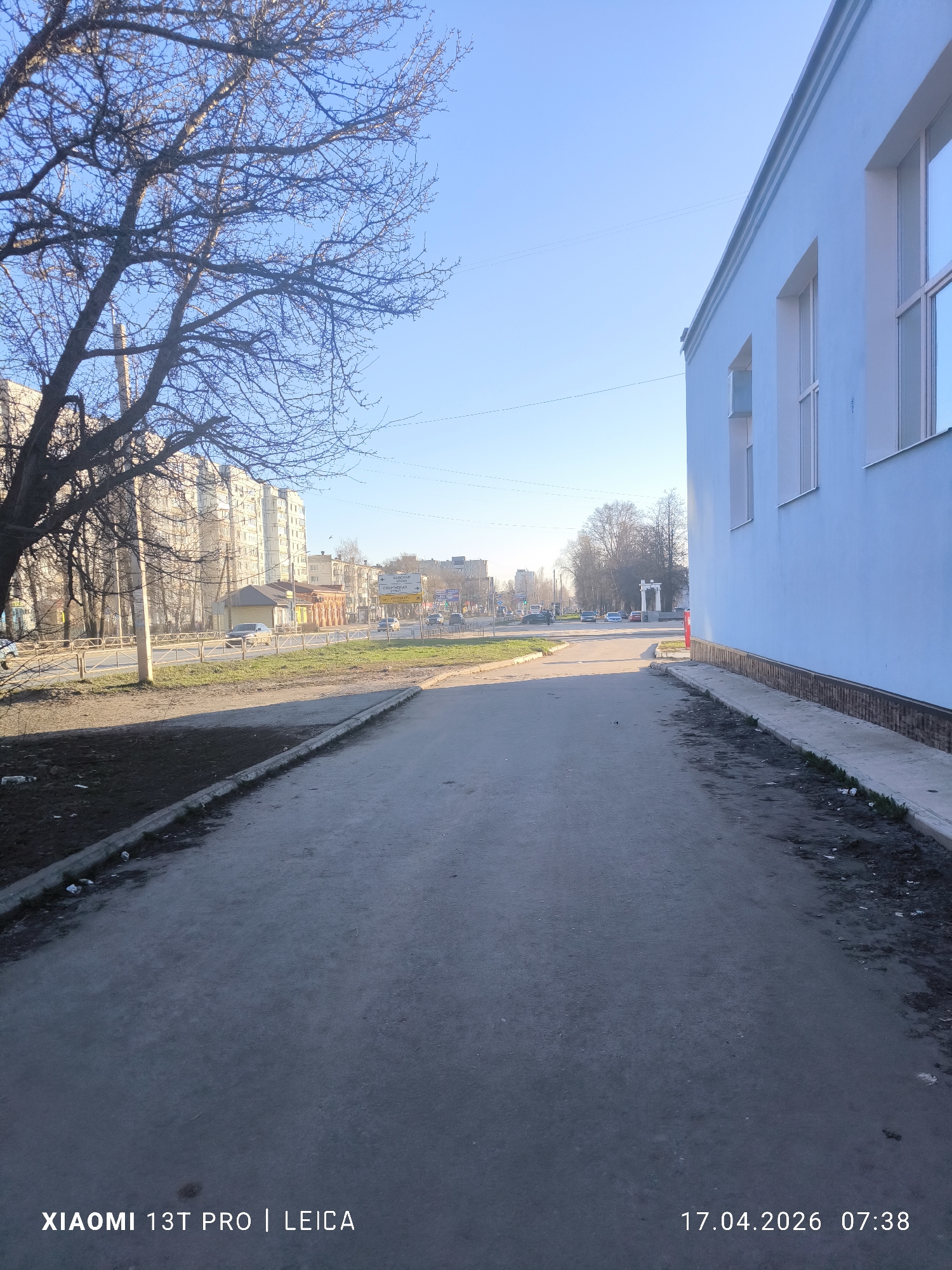 The image captures a quiet morning street scene in Chaikovsky, Russia, on April 17, 2026, at 07:38, under a clear blue sky. The weather appears sunny and calm, with bare trees suggesting late winter or early spring.
In the foreground, an asphalt path leads into the distance, bordered on the right by the light blue wall of a building with white-framed windows. The path's edges are somewhat unkempt, with patches of dirt and a visible plastic bottle near the curb on the left.
To the left, a large deciduous tree, currently devoid of leaves, dominates the scene, its branches extending into the clear sky. Beyond the tree, residential high-rise buildings are visible in the background.
In the mid-ground, a road runs parallel to the path, separated by a low metal fence and a strip of grass. Several cars are on the road, with some appearing to be in motion and others parked. A yellow street sign with Cyrillic text is prominent, clearly displaying "ЧАЙКОВСКИЙ" (Chaikovsky) at the top, confirming the location. Below it, "ЮНОСТИ" (Yunosti - Youth) is discernible, likely indicating a street name or direction. Smaller commercial-style buildings are visible behind the road and fence.
Further in the distance, a distinctive white archway or monument can be seen to the right of the road, adding a unique architectural element to the urban landscape. The overall setting is an urban environment, likely a residential or mixed-use area.
Visible text on the image watermark includes "XIAOMI 13T PRO | LEICA" and "17.04.2026 07:38". No people are visible in the frame, contributing to the peaceful atmosphere of the early morning.