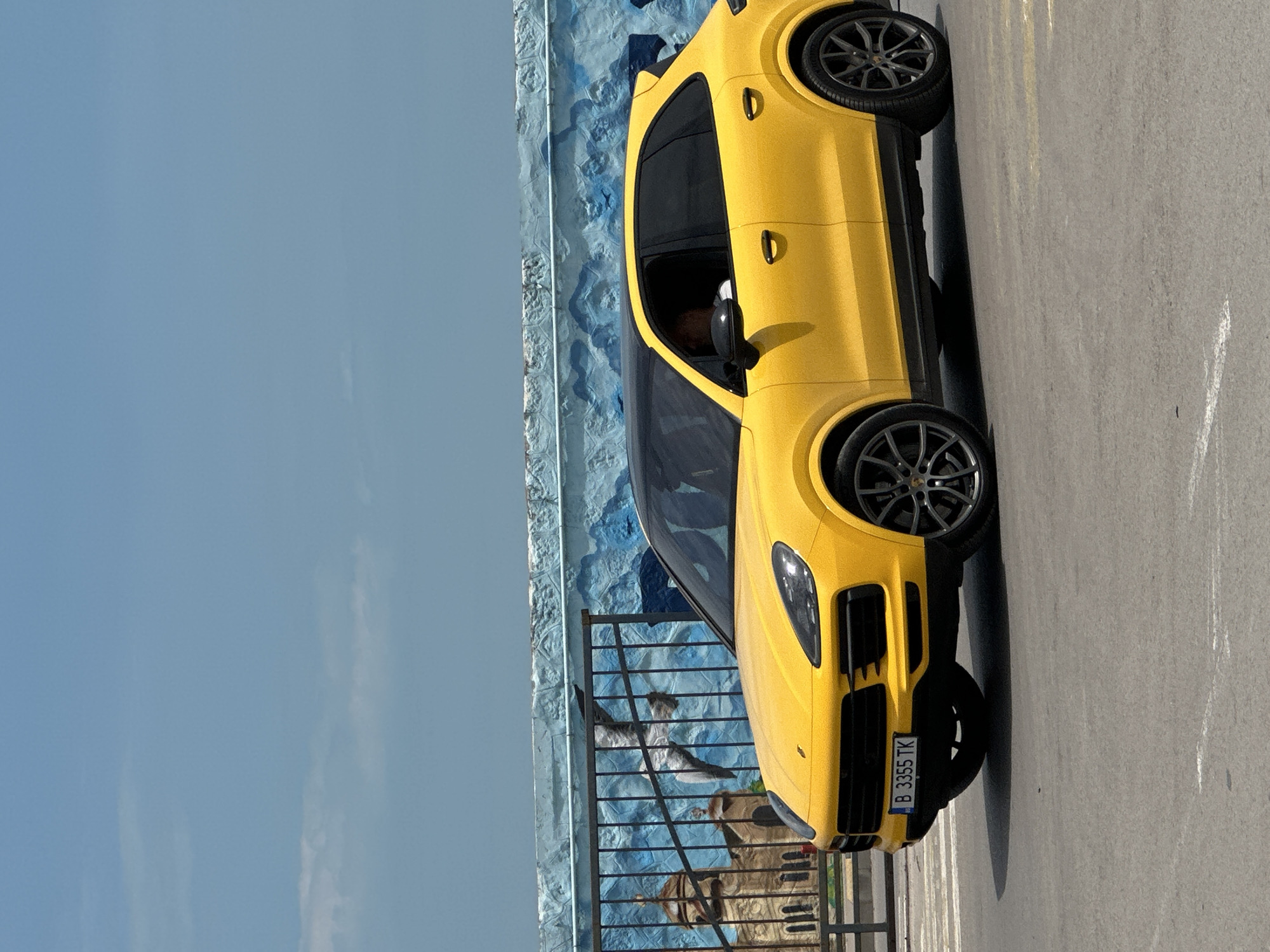 A yellow Porsche Macan is parked on asphalt. The car's license plate reads "B 3355 TK." A black metal fence with artwork in the background is behind the vehicle. In the background, there is a blue, slightly cloudy sky. Based on the license plate and other elements in the scene, this is likely in Varna, Bulgaria.
