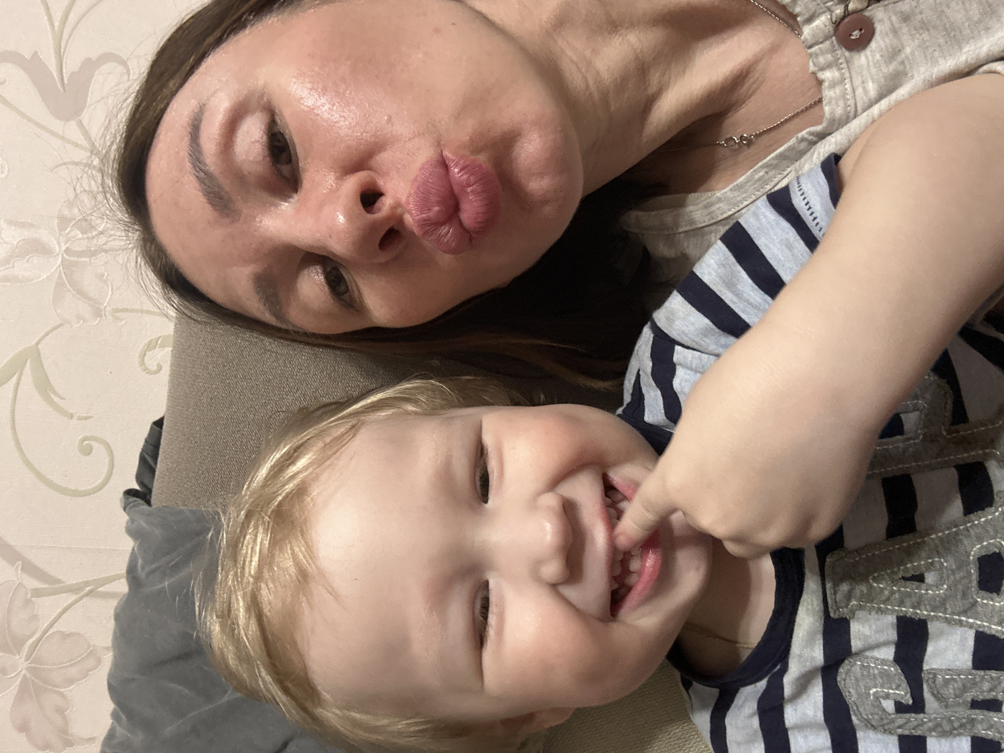 A woman and a young child are lying close to each other on what appears to be a bed or soft surface. The woman is on top with her head turned to the camera, making a pouting face. She has brown hair and is wearing a light-colored top with a button and a necklace. The child is below her, also turned towards the camera, smiling and touching their teeth with their fingers. The child has blonde hair and is wearing a striped shirt. The background shows a patterned wall. The image does not contain information about Votkinsk, Russia.