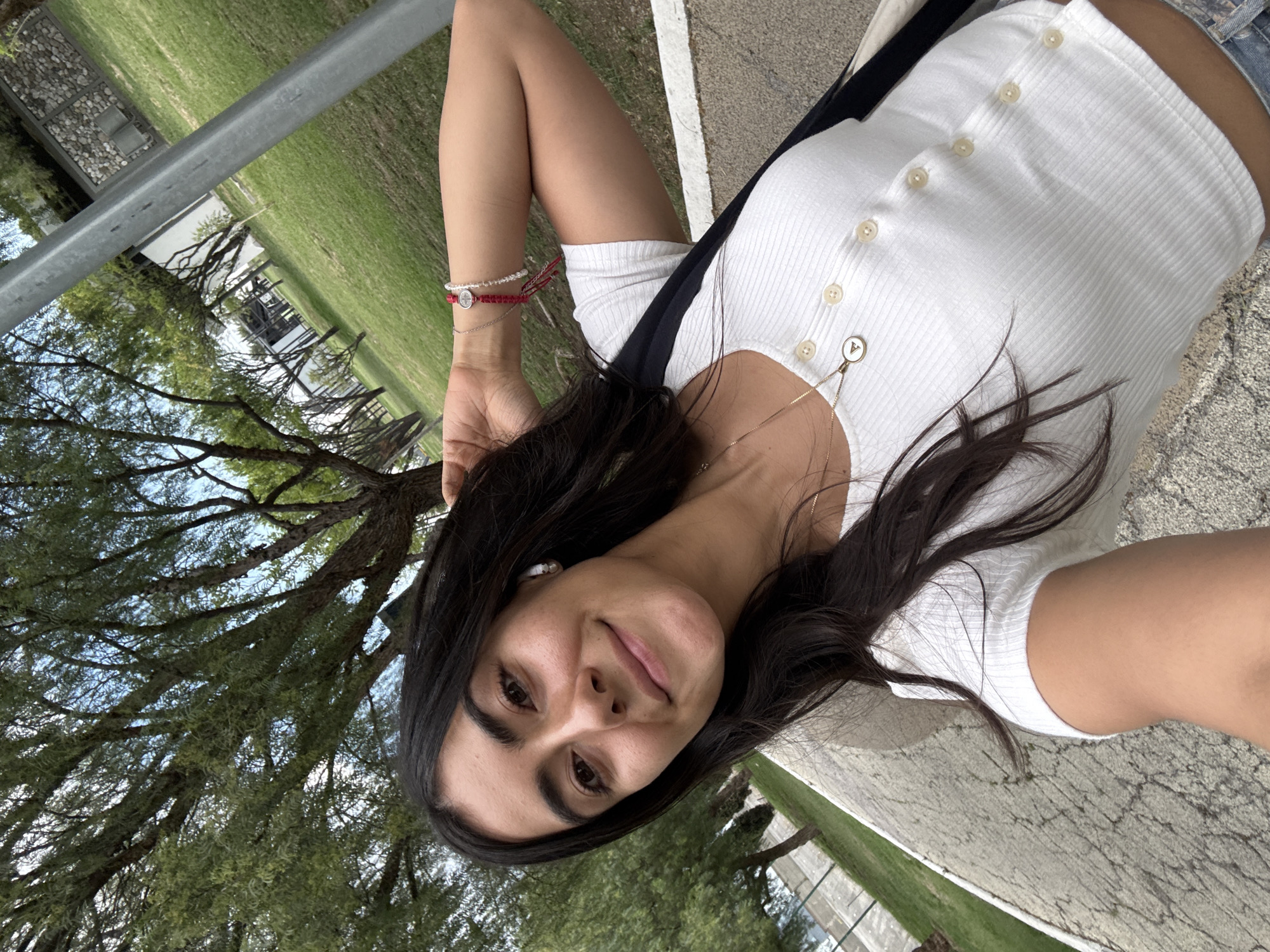A young woman is photographed outdoors at an angle. She wears a white, short-sleeved button-down shirt and has dark hair. She is smiling slightly and has bracelets on her left wrist. Behind her, there is a view of trees and grass, with a metal rail visible in the upper left corner. The ground appears to be cracked asphalt. The photo was taken at an angle. I have no information to determine the location of this photograph as being in San Bernardino Tlaxcalancingo, Mexico.