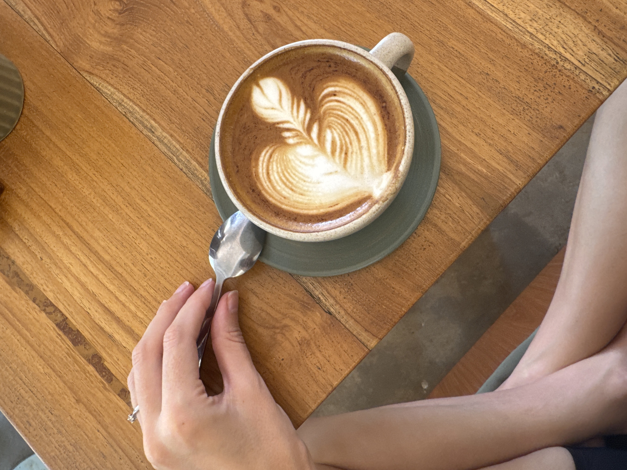 A cup of coffee with latte art, a spoon, and a person's hand and legs are visible on a wooden table. The coffee is in a ceramic cup, set on a green saucer. The latte art in the coffee appears to be a heart shape with a feathered design. The silver spoon is positioned to the left of the cup. A hand, likely belonging to the person, is holding the spoon. The legs of the person are also visible, crossed near the edge of the table. The table is wooden with a visible grain pattern. The location is unknown.