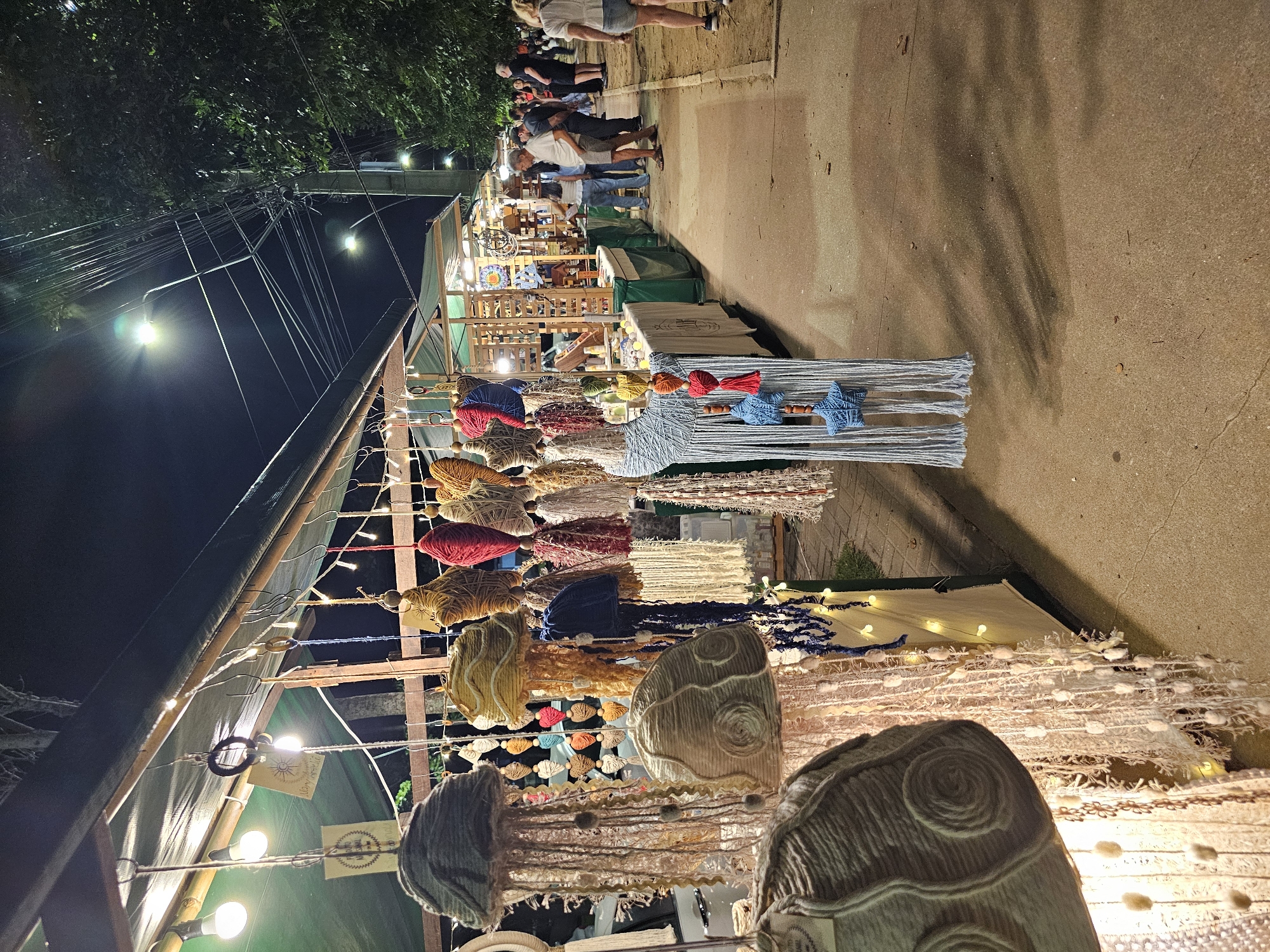 Captured at night in Armação dos Búzios, Brazil, the image presents a vibrant street market scene. A variety of handcrafted goods hang from wooden frames and display tables. Items include colorful, jellyfish-shaped decorations, tassel-like ornaments, and star-shaped accents. Patrons stroll along the side of the road, viewing the merchandise. Streetlights illuminate the scene. Overhead, a network of power lines stretches across the frame. The road surface is visible along the side of the frame.