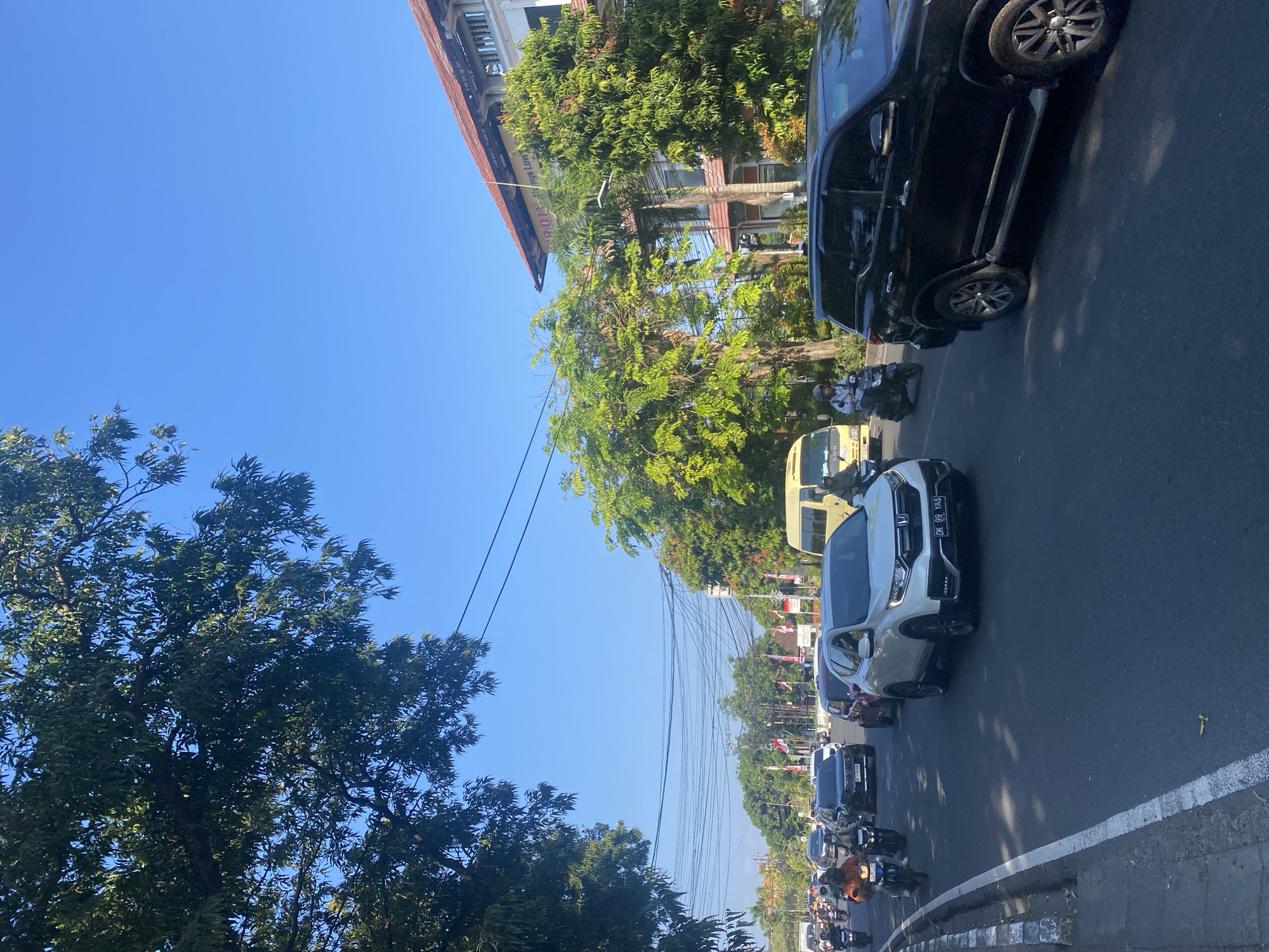This is a street-level view of moderate traffic on a sunny day in Denpasar, Indonesia. The road is paved with asphalt and has clear lane markings. On the left side of the road, there is a sidewalk with a curb. Trees with lush green foliage line the street, casting shadows on the road. In the background, there is a multi-story building with a red tiled roof and windows. Several vehicles, including cars, motorcycles, and a yellow van, are present on the road. People are seen riding motorcycles, some wearing helmets and casual clothing. The sky is clear and blue. The overall scene depicts a typical urban environment in Denpasar.
