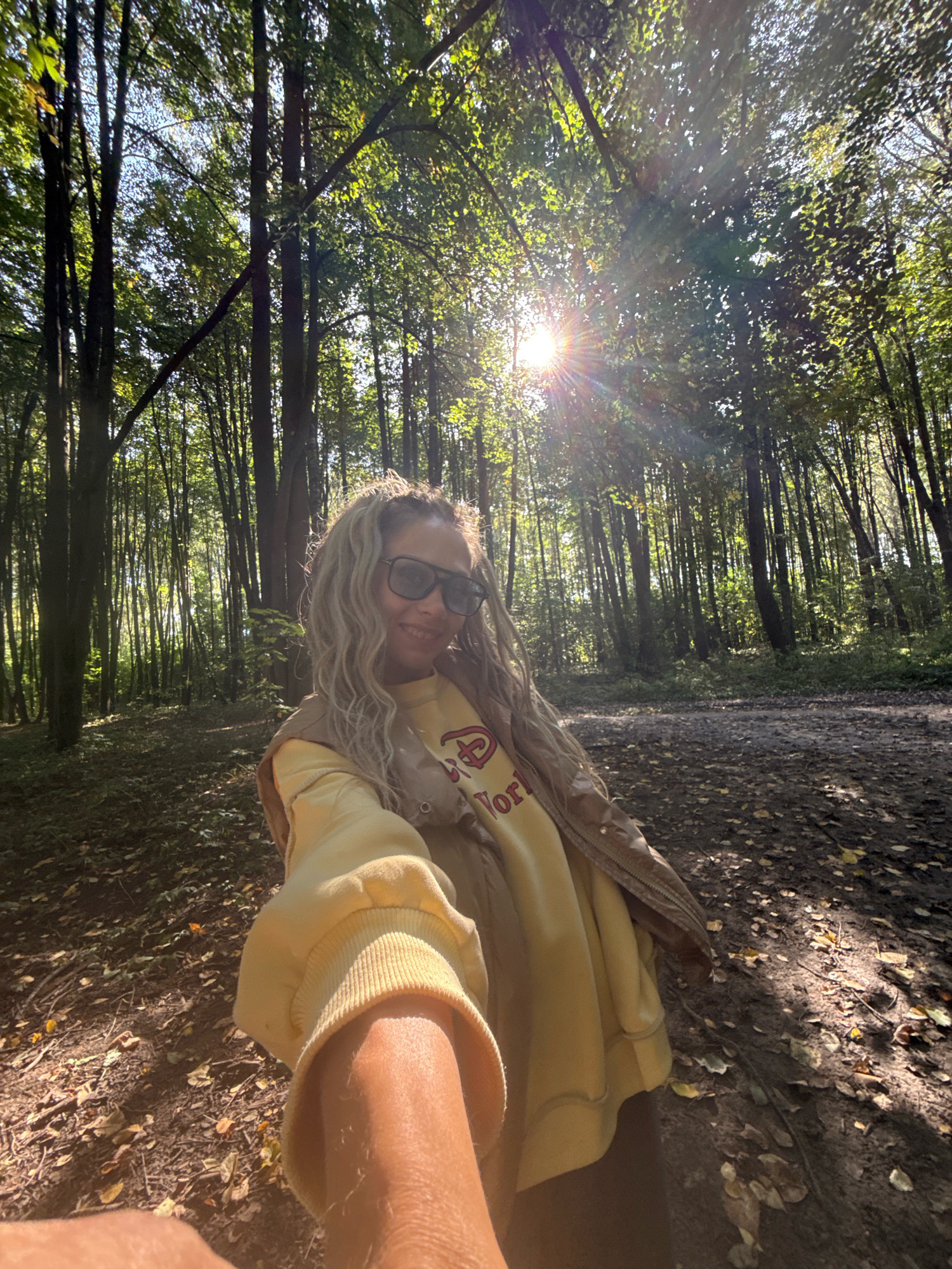 The image depicts a young woman taking a selfie in a forest. She has light-colored curly hair and is wearing sunglasses, a yellow sweatshirt, and a brown vest. She is smiling at the camera. The background shows a forest with tall trees, sunlight filtering through the leaves, and fallen leaves on the ground. The location is specified as the city of Rusanovo, Russia. The scene appears to be a sunny day, likely in autumn given the fallen leaves.