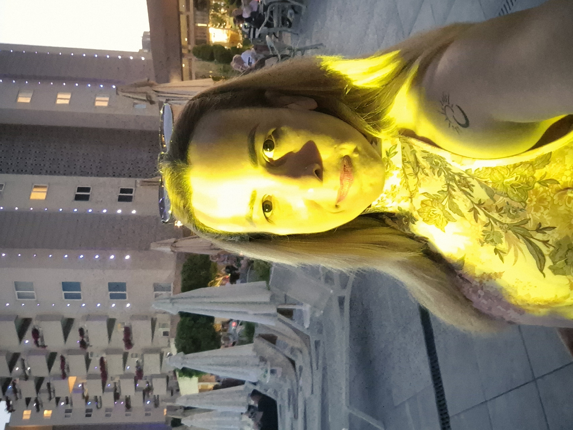 This media file is an image featuring a close-up of a woman's face, illuminated by a strong yellow light, which gives the scene a surreal and warm glow. The woman has blonde hair that falls around her shoulders and face. Her eyes are open and looking directly at the camera. She is wearing makeup, including lipstick that appears to be a reddish-pink shade. On her upper arm, a small, dark tattoo resembling a sun or an abstract circular symbol is visible. She is also wearing a yellow garment with a floral pattern.
The background suggests an outdoor setting, possibly a patio or dining area. There are hints of buildings with windows and lights in the distance, as well as what appear to be outdoor tables and chairs, some covered with white fabric, possibly awnings or umbrellas. In the further background, there are blurred figures of people and some greenery, indicating a public space or gathering.
The overall impression of the image is informal, perhaps a selfie. The strong lighting creates a dramatic effect, washing out some details of the environment and emphasizing the woman's face. The time of day is unclear due to the artificial lighting, but the faint sky in the top left corner could suggest either dawn, dusk, or artificial lighting mimicking daylight. There is no visible text in the image.