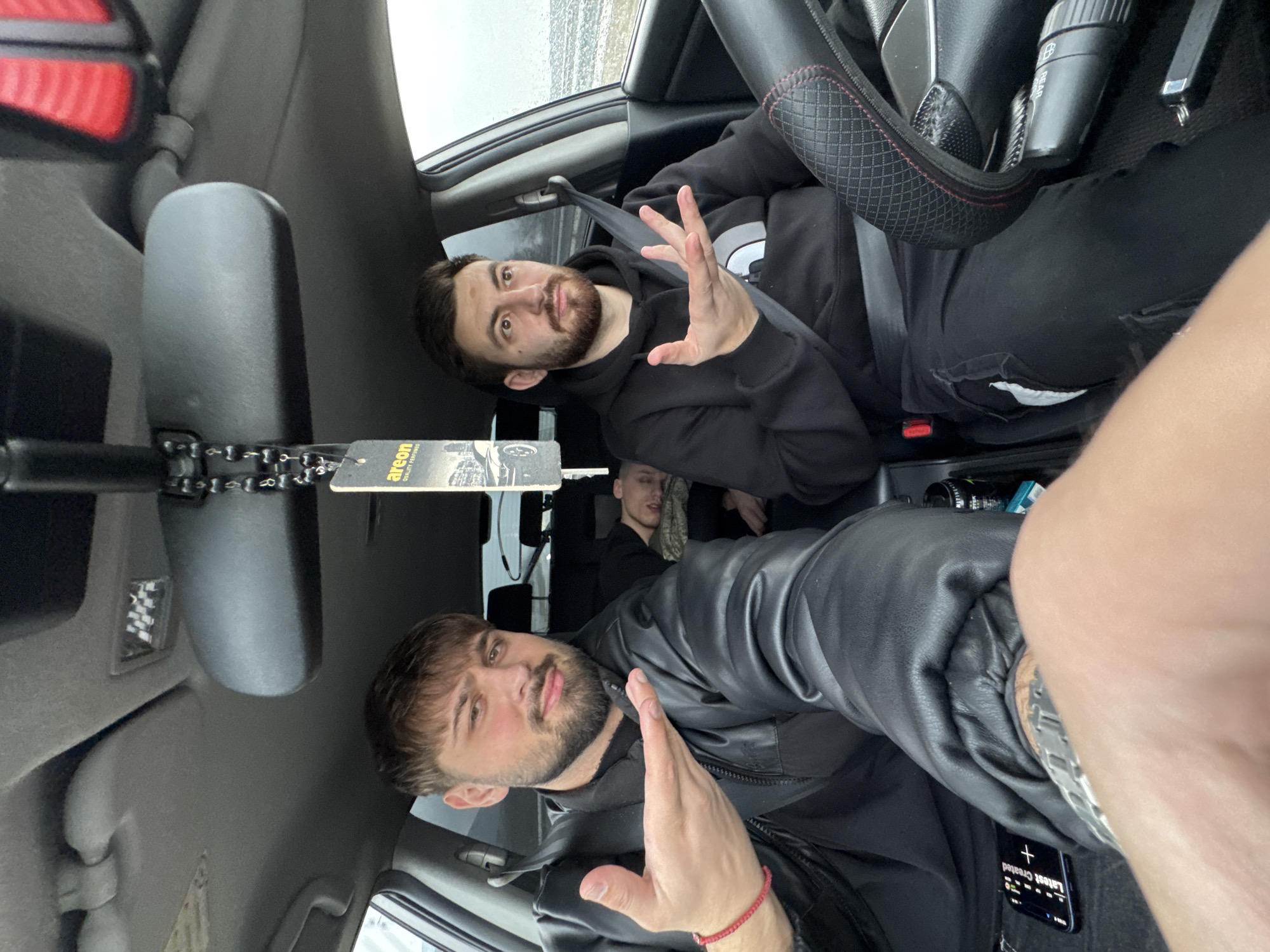 The image displays the interior of a passenger vehicle, captured from a low, front-facing perspective, likely a selfie. Three adult males are present.
The individual in the driver's seat (right side) is a male with dark hair and a beard, wearing a black hooded sweatshirt with a white logo and dark trousers. He is secured by a seatbelt. His left hand is raised with fingers splayed, mid-gesture, while his right hand rests on a black steering wheel featuring a red-stitched, diamond-patterned cover. His expression suggests a questioning or confused demeanor.
The individual in the front passenger seat (left side) is a male with dark hair and a beard, wearing a black leather jacket over a dark garment. A red bracelet is on his right wrist, and a silver-toned watch is on his left wrist. His right hand is extended with the palm open, in a gesturing motion, and he exhibits a slight smirk. A smartphone, displaying "Latest Created," is visible on the center console or floor below him.
A third male is visible in the rear seat, appearing to be asleep or resting, with his head tilted back and to the right. He wears a dark top and what appears to be a light-colored or patterned scarf around his neck.
The vehicle's interior features a grey headliner, a rearview mirror, and an overhead light with a checkered pattern. A black beaded chain with an "AREON quality perfume" air freshener hangs from the rearview mirror. Through the vehicle's windows, a blurred, overcast exterior environment is visible, lacking specific landmarks. The precise location of the vehicle within the city of Nevsha, Bulgaria, cannot be visually confirmed from the image provided.