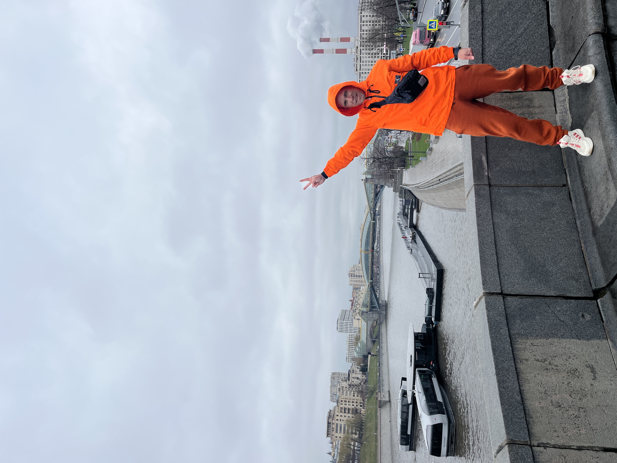 A man wearing an orange hooded sweatshirt, orange sweatpants, and white sneakers stands on a concrete ledge, possibly a bridge or embankment, with his left arm extended, making a peace sign with his fingers. He is wearing a black fanny pack. The background shows a cityscape with a river, possibly the Moskva River, featuring boats and a bridge in the distance. The sky is overcast. In the far background, there are industrial chimneys emitting smoke. The location appears to be in Moscow, Russia.