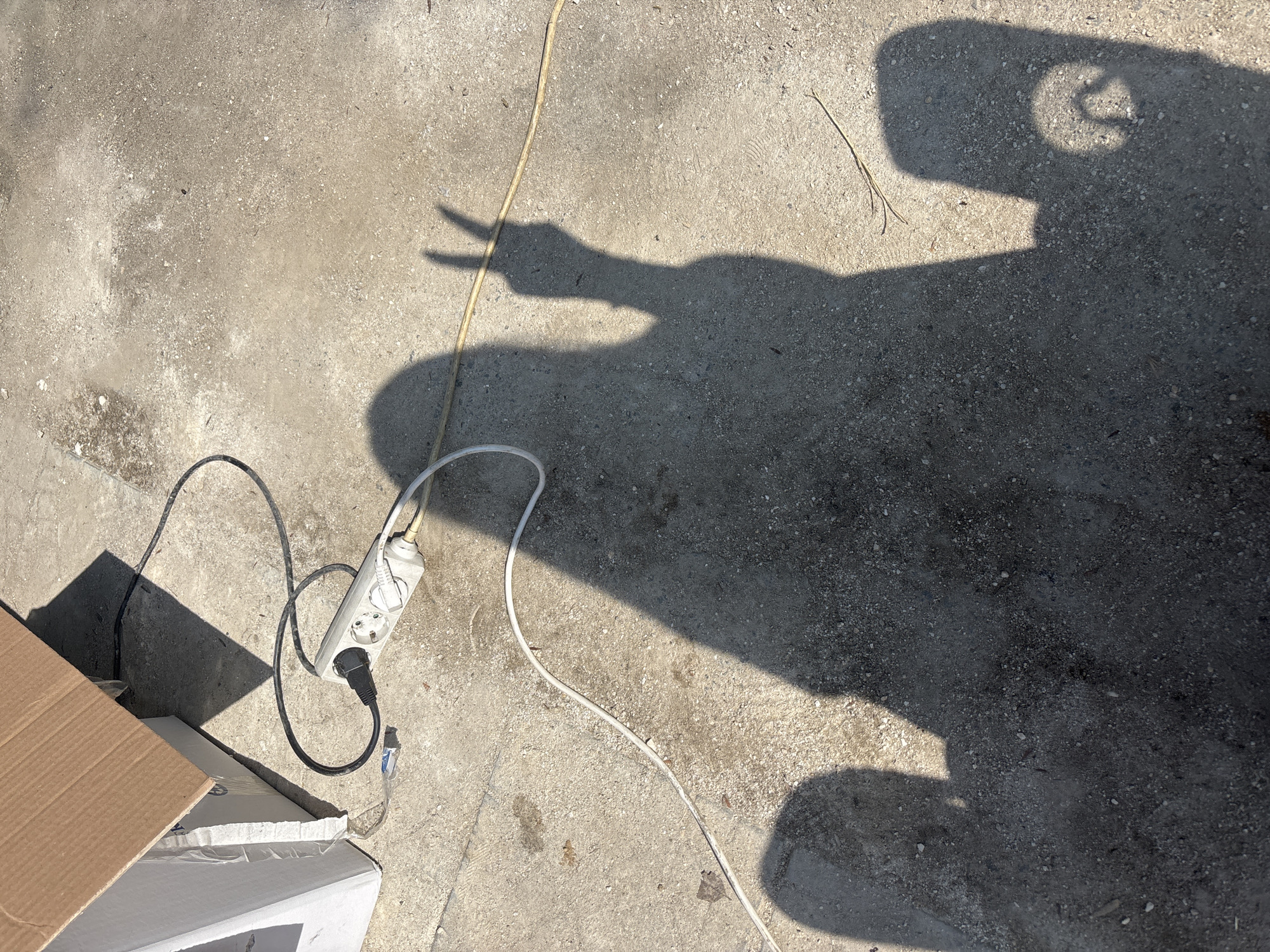 The scene is an outdoor ground area in Kostievo, Bulgaria, illuminated by strong direct sunlight, casting distinct shadows. The ground surface is composed of light gray, speckled concrete or compacted gravel, exhibiting variations in tone and minor debris. In the upper left portion of the image, a partially open brown cardboard box is positioned next to a white, box-like container. Centrally located, a white multi-socket power strip lies on the ground. A black electrical cable, forming a loose loop, is plugged into one socket and extends towards the boxes. A white electrical cable is plugged into another socket and extends diagonally downwards and to the left. A yellowish electrical cable is visible extending from the right side of the power strip towards the lower right. Multiple human shadows are cast across the ground. A prominent shadow, originating from the right and extending across the center, depicts a person with their right hand raised, casting a clear "peace sign" (index and middle fingers extended in a V-shape). To the lower left of this primary shadow, a partial shadow of a head and upper torso is visible. A third, partial, rounded shadow appears in the lower right, consistent with another human presence or an object held in hand.