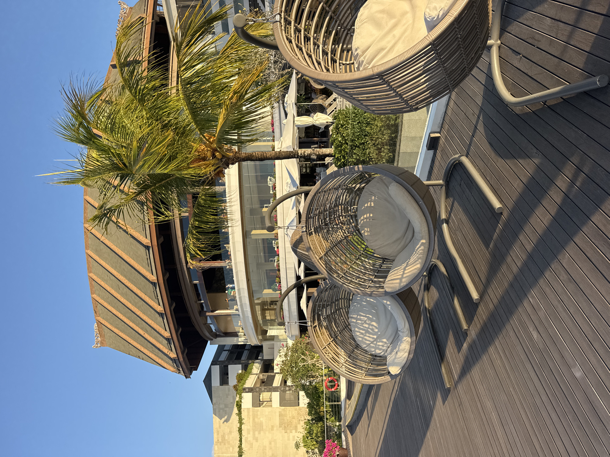 The image depicts an outdoor seating area with three hanging egg chairs on a wooden deck. The chairs are made of woven material and have light-colored cushions. In the background, there is a multi-story building with a distinctive roof, a palm tree, and lush greenery. The scene is bathed in warm sunlight, suggesting it is either morning or late afternoon. The location appears to be a resort or a public space in Kuta Selatan, Indonesia, known for its beach resorts and tourist attractions. The sky is clear and blue.