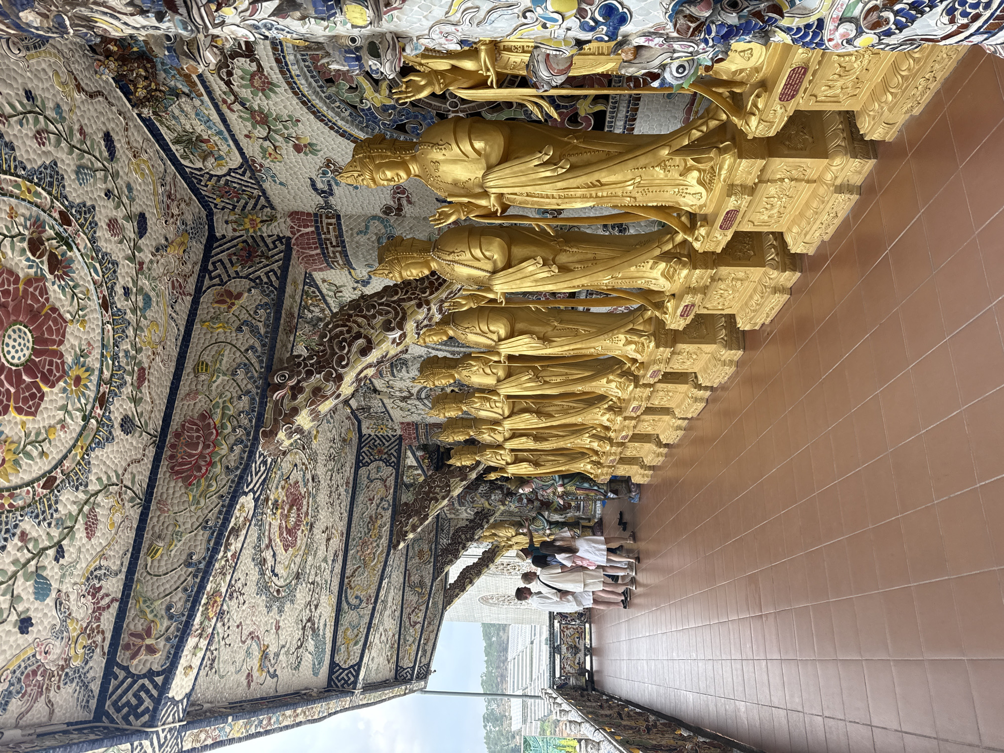 The image depicts a view inside a building in Xuân Thành, Vietnam. A row of golden statues of figures, possibly deities, are mounted on a wall above eye level. The ceiling and wall to the left are decorated with intricate mosaic tile patterns featuring floral motifs and geometric designs. Several people are walking along a tiled floor, looking at the statues. Outside, there is a view of a bright sky, possibly a distant landscape.