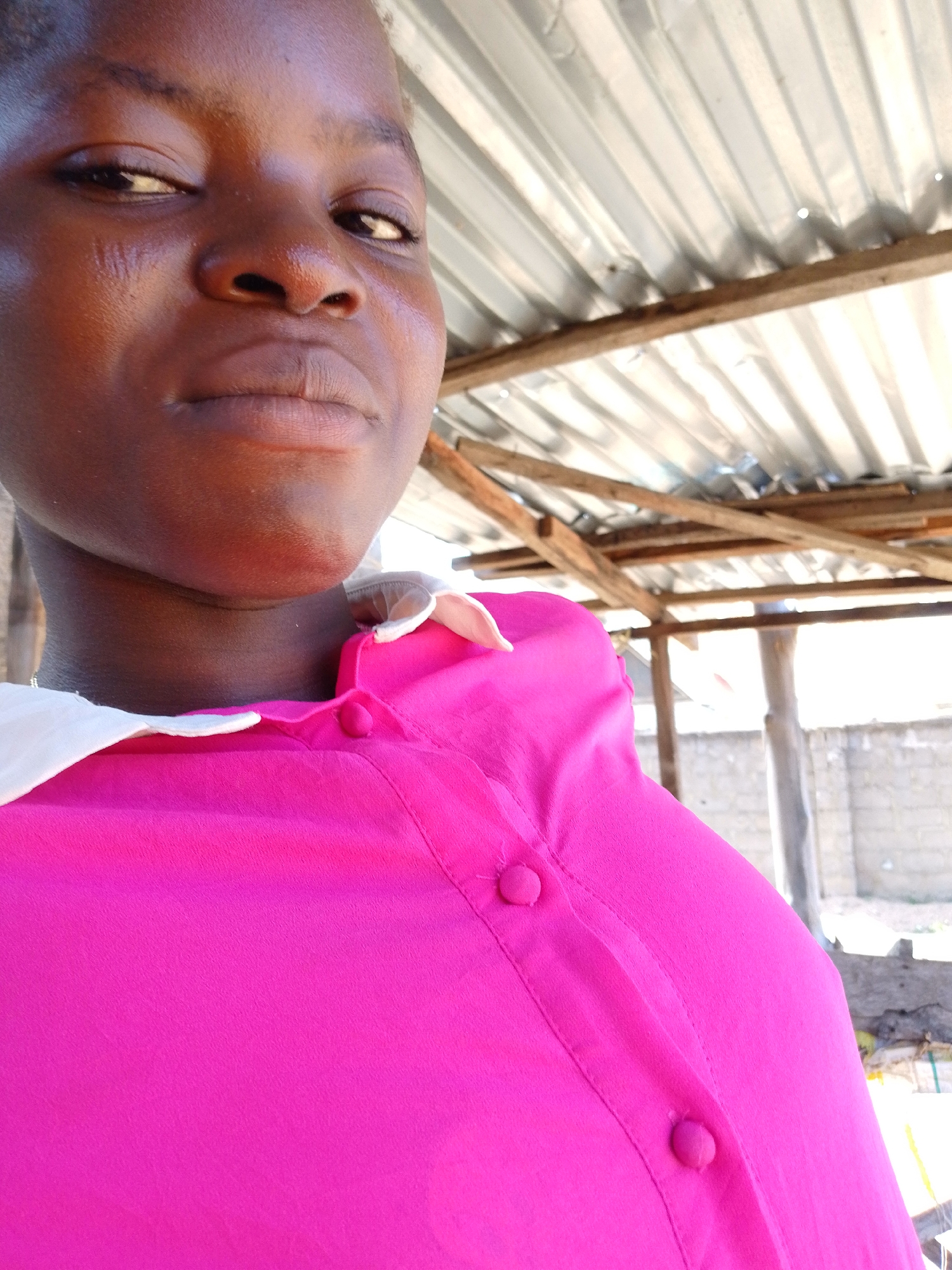 A close-up, slightly angled shot features a dark-skinned woman with a serious expression. She is wearing a bright pink, button-down shirt with a white collar. The camera angle is from below, looking upwards towards her face.
The background reveals a partially covered outdoor area. Corrugated metal roofing is visible, supported by wooden beams. In the distance, there is a glimpse of a light-colored building or wall.
Given the context of Lokoja, Nigeria, it's likely an outdoor or semi-outdoor area providing shade. The specific location within Lokoja cannot be determined from the image alone.