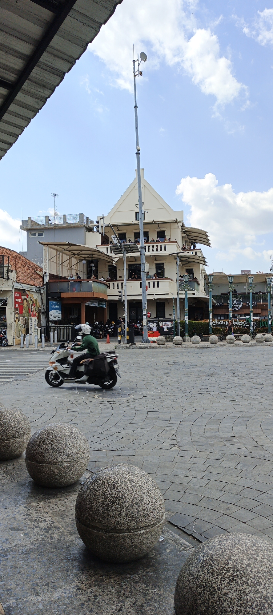 In Yogyakarta, Indonesia, a motorcycle rider wearing a green shirt and a white helmet is captured in motion on a cobblestone street. The rider, with a black bag attached to the rear of the motorcycle, is moving from left to right. In the background, a multi-story building with a distinct white facade and balconies is visible, suggesting an urban setting with commercial or residential spaces. The sky is blue with scattered white clouds. The foreground features large, round, textured stone spheres arranged on the cobblestone pavement, possibly serving as bollards or decorative elements. The overall scene depicts a street in Yogyakarta on a clear day.
