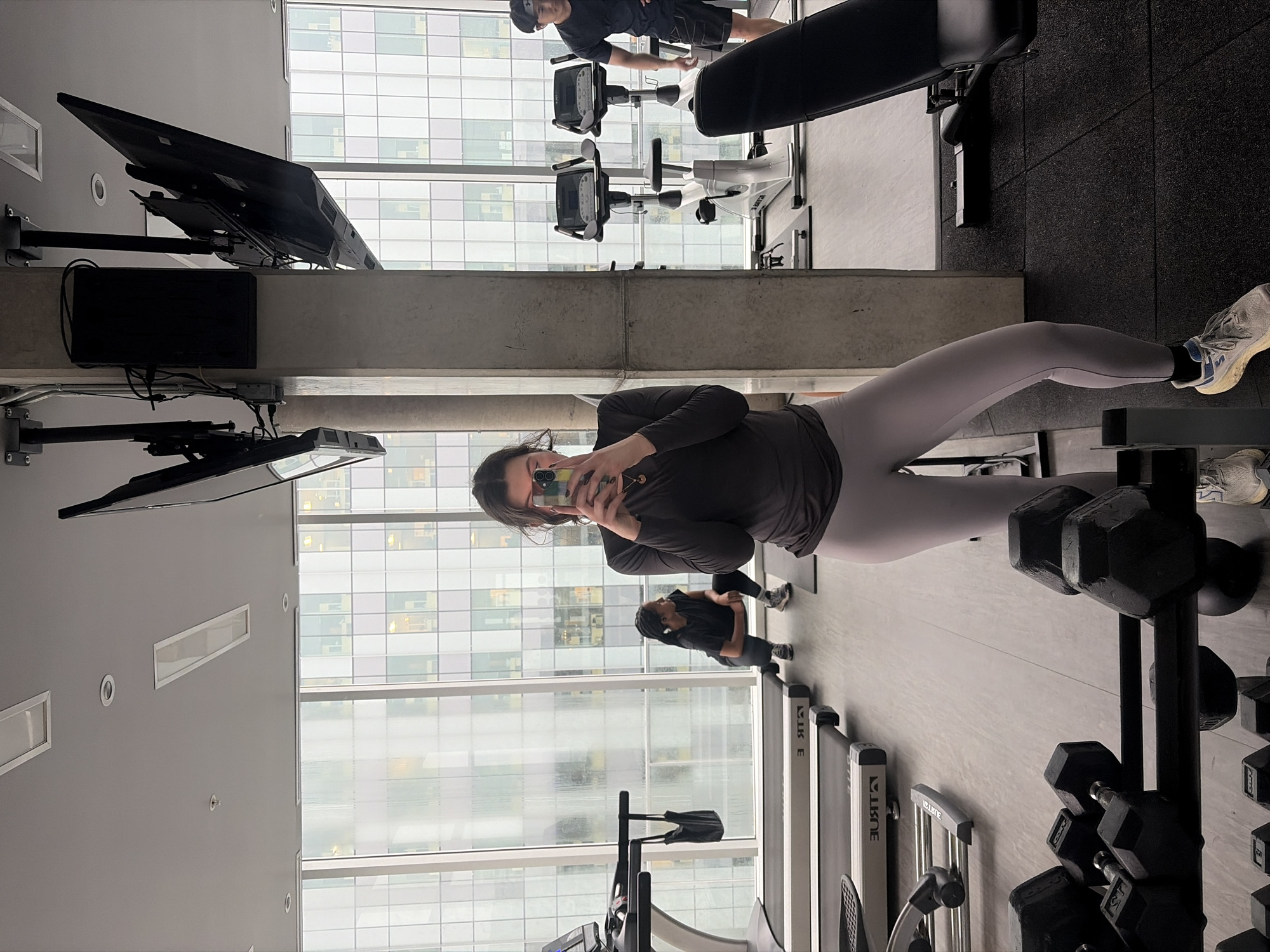 This image captures an interior view of a modern gym or fitness center, primarily featuring a woman taking a mirror selfie. The woman, positioned centrally, wears a dark long-sleeved top, light grey or lavender leggings, and white and grey athletic shoes. She has dark hair, wears glasses, and is holding a smartphone with a colorful case to take the picture.
The setting is a well-lit gym with large, grid-patterned windows that reveal an urban landscape of other tall buildings, consistent with a high-rise location in a city like Montreal. The natural light suggests it is daytime, with an overcast or cloudy sky.
Various gym equipment is visible throughout the space. In the foreground, a rack of black hexagonal dumbbells stands next to the main subject. Two treadmills, bearing the brand name "TRUE" on their sides, are visible in the mid-ground. Further into the gym, two stationary bikes are present, with one occupied by a person in a dark t-shirt and shorts. Another individual in dark athletic wear stands near a treadmill to the left of the main subject. A weight bench is also discernible.
The gym's design is contemporary, featuring light-colored flooring, grey walls, and mounted black flat-screen monitors. Recessed lighting and rectangular fixtures are integrated into the ceiling. The activity is primarily a casual selfie, with other individuals engaged in exercise in the background.