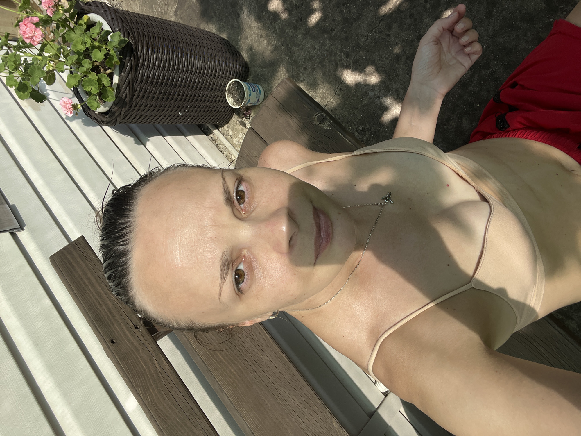 A woman is sitting on a wooden bench outdoors, with a white vinyl-sided wall behind her. She is looking at the camera. She is wearing a light-colored camisole and red shorts. To her right, a dark wicker planter holds pink flowers. A small white cup with soil is also on the ground next to the planter. The ground is concrete and shaded by dappled sunlight.
