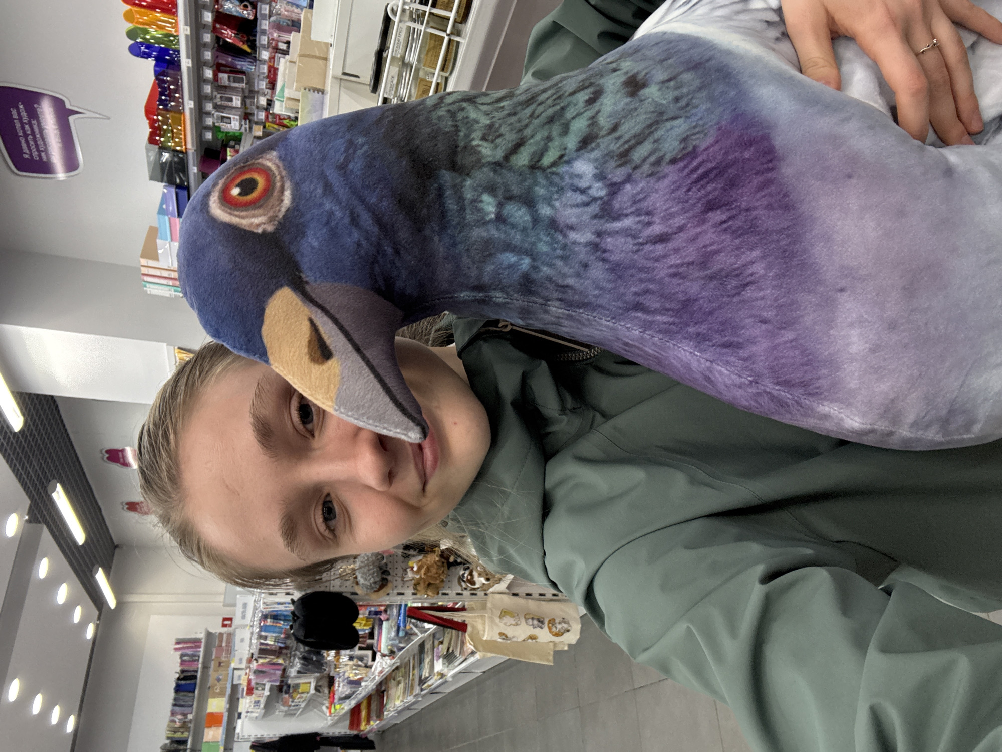 A fair-skinned woman with blonde hair, wearing a green jacket, holds a large plush pigeon in a store. The pigeon has a detailed, fabric print simulating feathers in shades of blue, purple, and green, with a tan beak and a printed eye. The woman is looking at the camera. The store has shelves stocked with various items, illuminated by overhead fluorescent lights. A store sign and colorful merchandise are visible in the background. The location is in the city of Izhevsk, Russia.