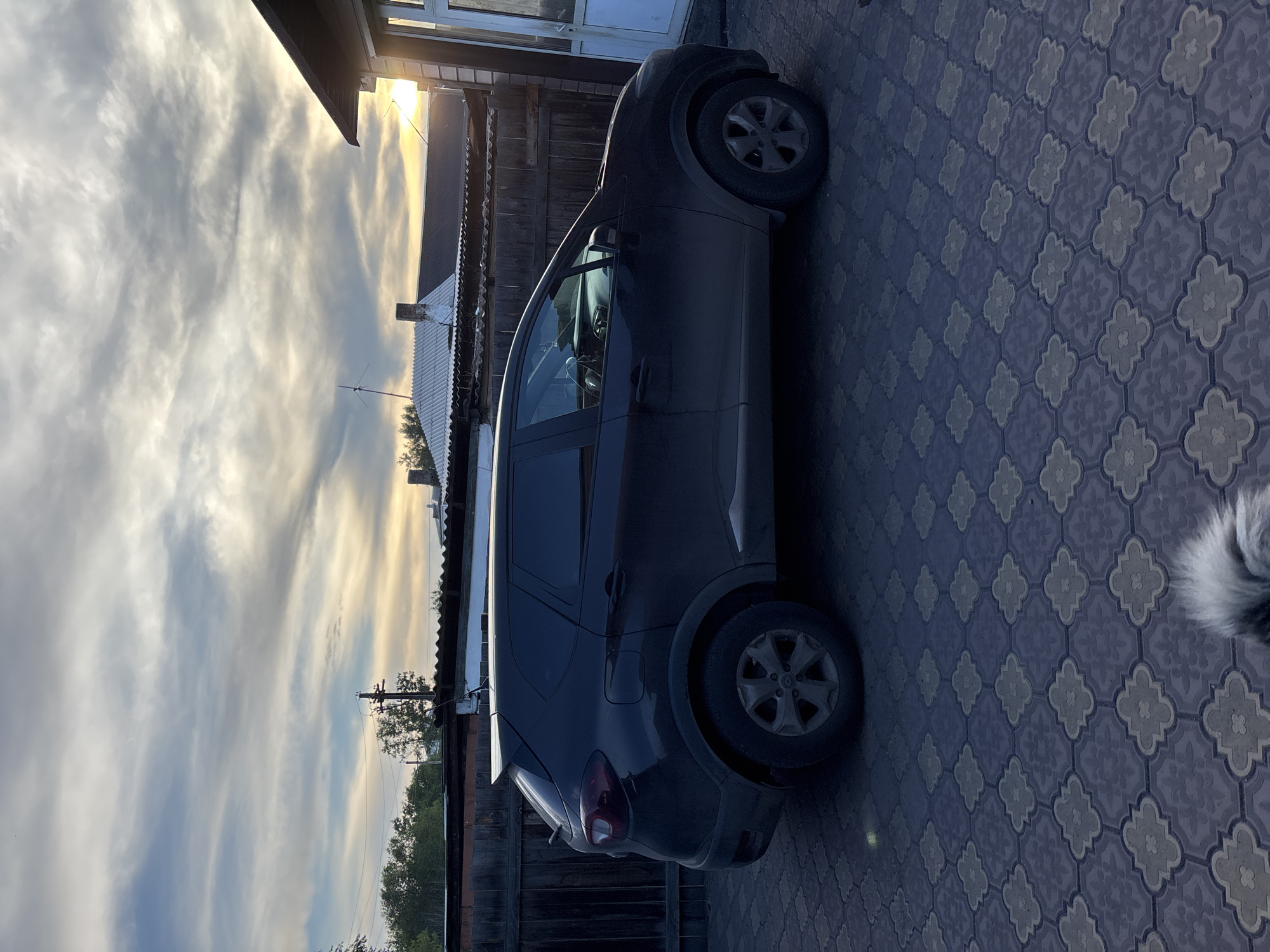 In Chernogorsk, Russia, at the side of a residential building, a dark colored, four-door crossover SUV is parked on a decorative patterned concrete driveway. The vehicle is facing right, with its rear slightly angled towards the viewer. The car appears dusty. Behind the vehicle, a wooden fence runs along the property line, with trees and structures visible beyond it. The sky above is cloudy with a mix of grey and white clouds, and the sun is setting, casting a warm glow. A portion of a building with a white door is visible on the right side of the frame. The lower portion of the image shows the patterned paving stones of the driveway.