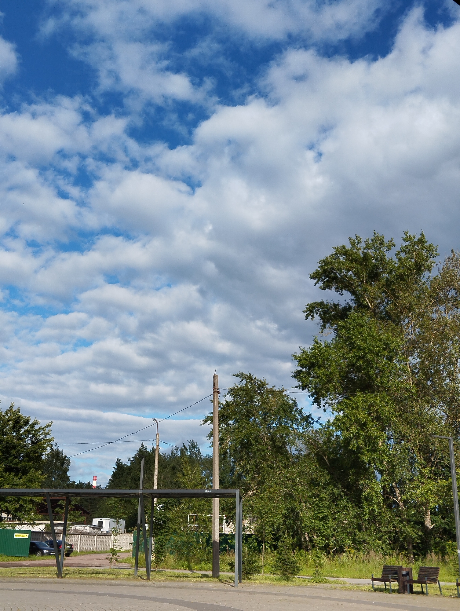 The image depicts an outdoor scene with a focus on trees and the sky. A tall, red and white striped industrial chimney is visible in the distance, to the left of center. The sky is blue with scattered white clouds, some of which are tinged with grey, suggesting varying atmospheric conditions. In the foreground, there is a paved area with a textured surface, possibly a walkway or plaza. To the right, two wooden benches with dark seats and metal frames are placed next to each other, with a small trash receptacle situated between them. Lush green trees with dense foliage dominate the middle ground and right side of the image. A metal structure, resembling a canopy or shelter with vertical and horizontal supports, is present in the middle ground. Behind this structure, there are buildings, including a white building and a fence made of concrete blocks. A dark-colored car is parked to the left of the structure, near a green corrugated metal structure. The overall impression is a somewhat urban or suburban park-like setting with industrial elements in the background. The location is identified as Kingisepp, Russia, which is a city in Leningrad Oblast.