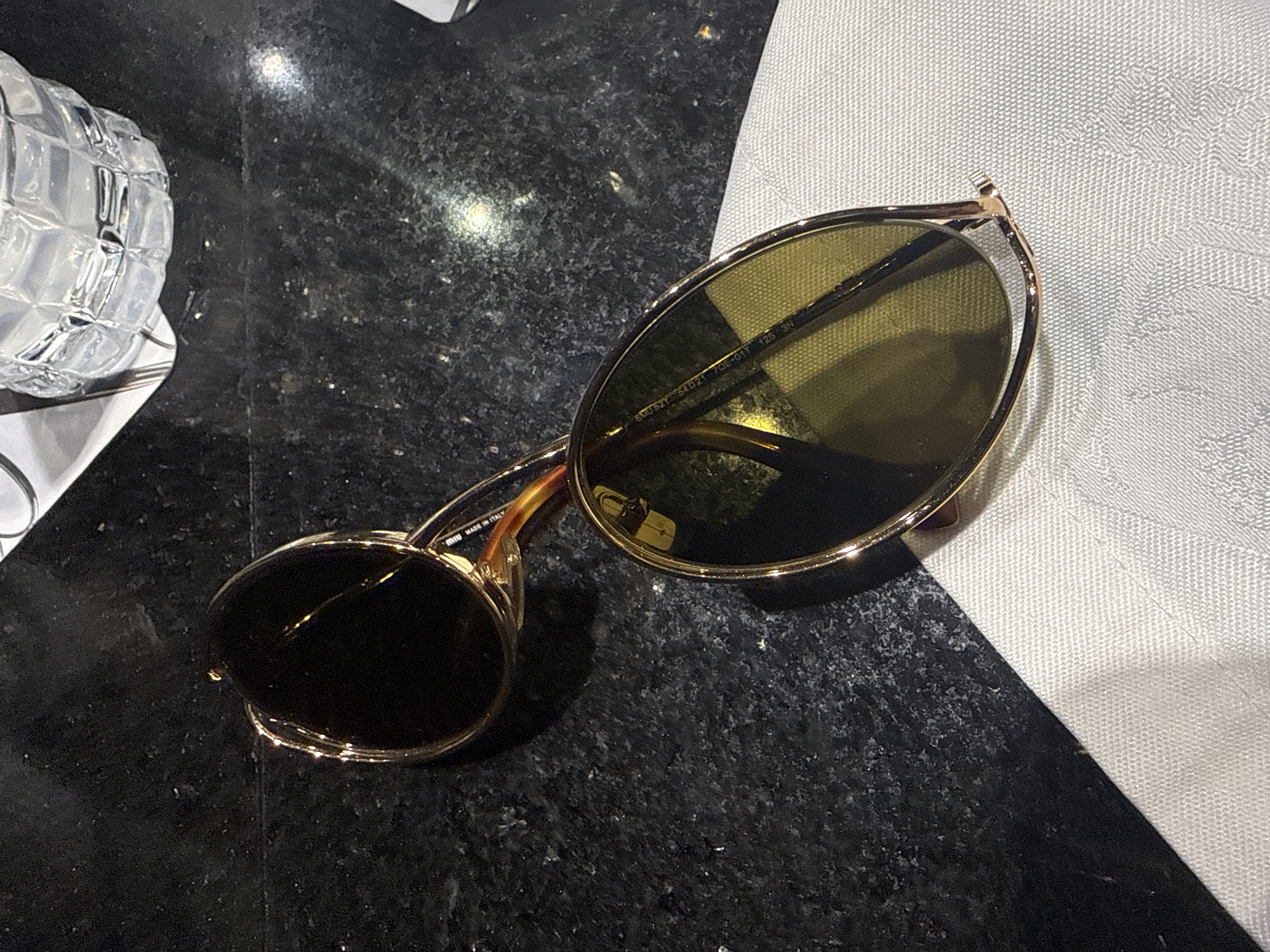 The image shows a pair of sunglasses on a black, speckled surface next to a white, textured cloth. The sunglasses have gold-colored frames and dark lenses. To the left, there is a clear, textured glass object. Given the items and the context, it's plausible that the location is a restaurant in the city of Puebla, Mexico. However, without further context it is not possible to verify that the scene is specifically set in Puebla.
