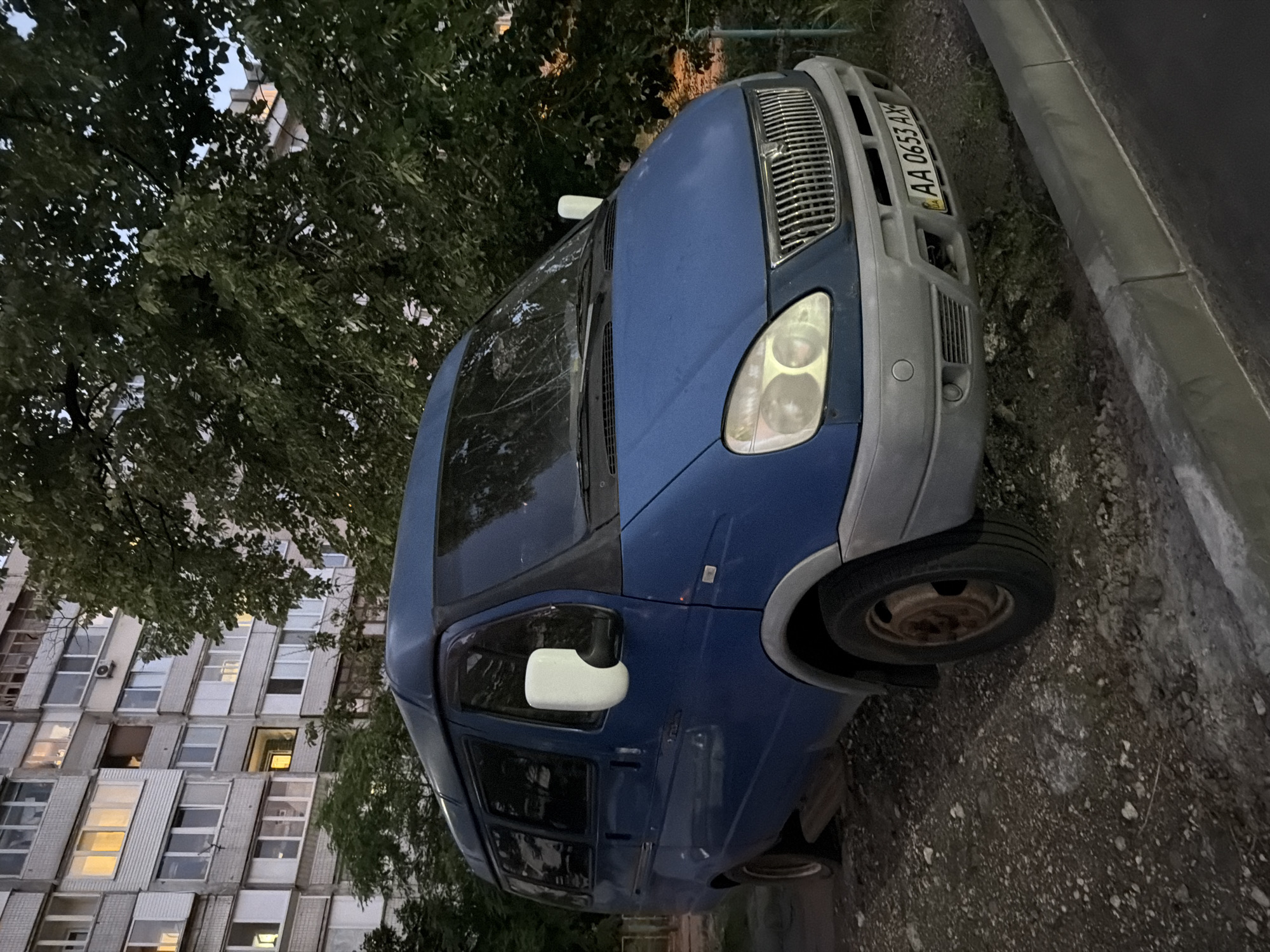 A blue Gazelle van is parked on a dirt and gravel lot next to a sidewalk. The van is positioned at an angle, facing slightly towards the viewer's right. It has tinted windows, a gray bumper, and a silver grille. The license plate is visible and reads "AA 0653 AX". Behind the van, a large, mature tree with dense green foliage obscures much of a multi-story apartment building. The building has numerous windows, some of which are lit, suggesting occupancy. The sky appears overcast. The ground is uneven and appears to be a mixture of dirt, gravel, and scattered small stones.