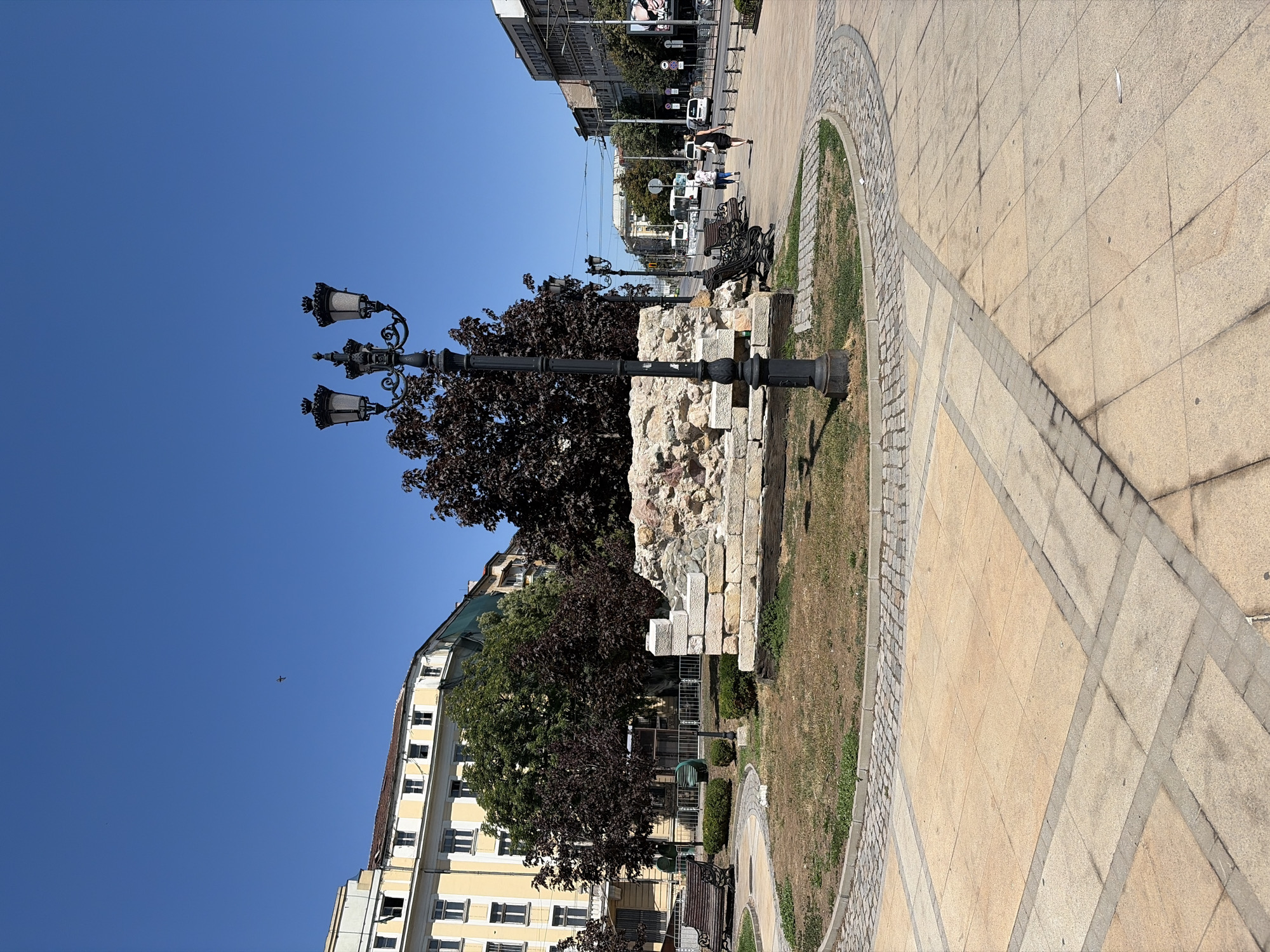 The image depicts a clear, sunny day in Sofia, Bulgaria, likely in a public square or park area. In the foreground, a paved area with large, light-colored square tiles is visible, bordered by a curved section of smaller cobblestones. A street lamp with three ornate black fixtures stands prominently in the mid-ground.
Behind the lamp is a circular grassy area with some brown patches, indicating dry conditions. At the center of this grassy area, a low-lying, rugged stone structure, appearing to be part of an ancient ruin or monument, is partially visible. Several dark-leaved trees are positioned around this structure and the grassy area.
To the left, a wooden park bench faces the grassy area. Beyond the bench, a light-yellow building with multiple windows and a tiled roof is seen. In the background, a street stretches into the distance, lined with buildings and trees. A few people are walking on the sidewalk of this street, and several vehicles, including a bus and cars, are present. The sky is a vibrant, cloudless blue, with a small bird or aircraft visible in the upper left quadrant.