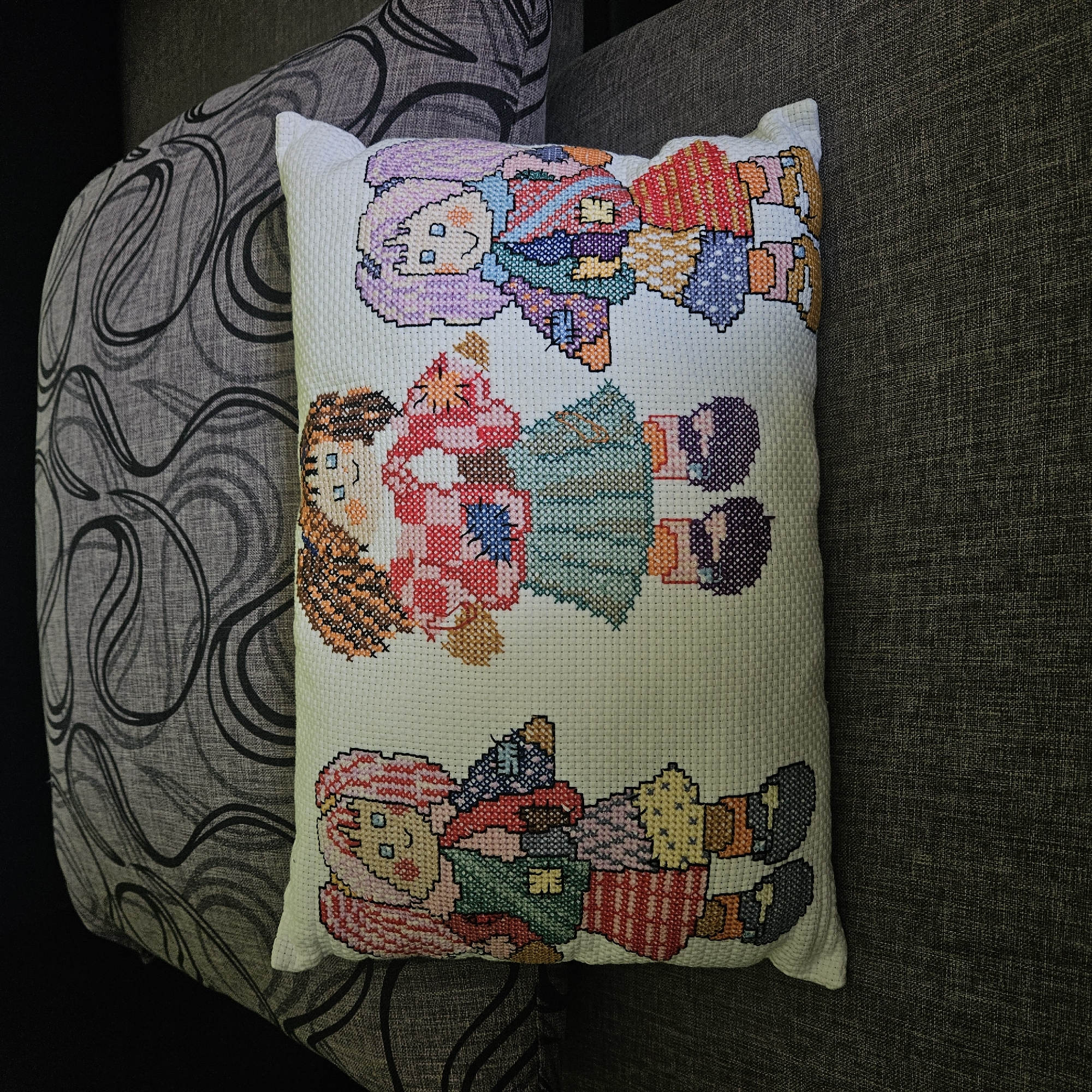 A close-up shot presents a rectangular throw pillow resting on a couch. The pillow is white and adorned with three cross-stitched figures of dolls. From top to bottom, the first doll has lilac hair and a colorful outfit. The second doll has orange-brown hair and a red and green outfit. The third doll has pink hair and a colorful outfit. The couch has two sections, one with a gray swirling pattern and the other with a dark gray, tightly woven fabric. There are no discernible indications to specify that this image was taken in the city of Stara Zagora, Bulgaria.