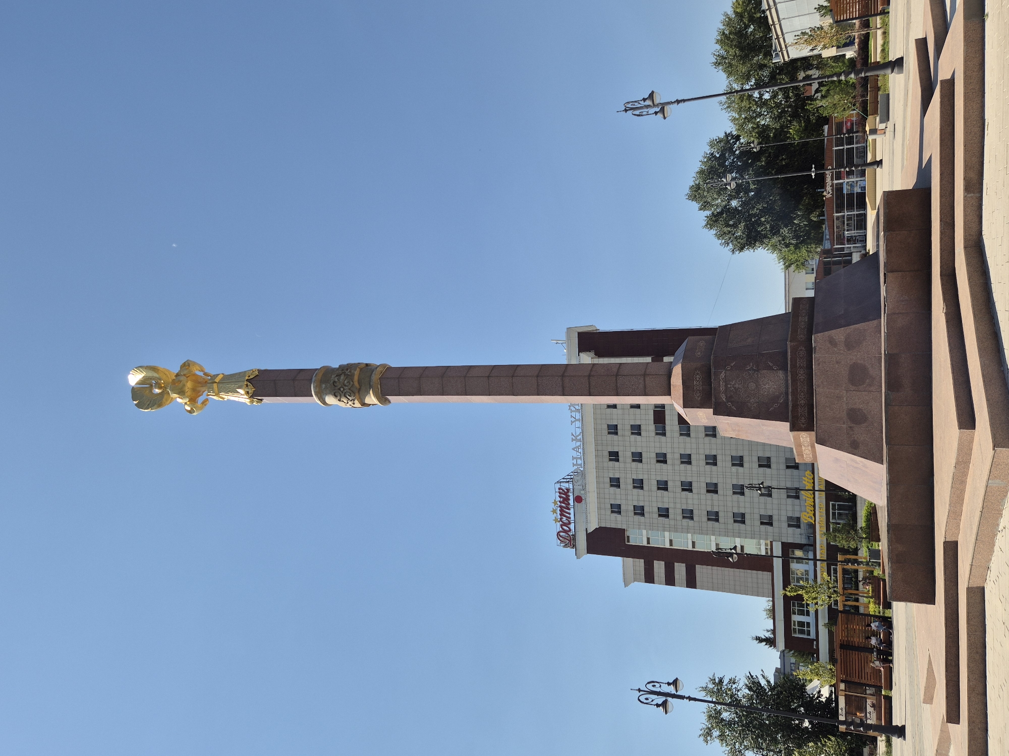 This is a ground-level, eye-level, wide-angle, outdoor photograph taken on a clear, sunny day in Kokshetau, Kazakhstan. The primary subject is a tall, bronze-colored monument topped with a golden eagle statue. The monument is made of granite and features intricate carvings and a wide, stepped base. In the background, a modern, multi-story hotel with a red sign reading "Достык" (Dostyk) and "Қазақстан" (Kazakhstan) is visible. To the left of the monument, a street lamp with two lights stands on the sidewalk. Several people are seated on benches in the middle ground, and trees and other buildings can be seen further in the background. The ground is paved with light-colored tiles.