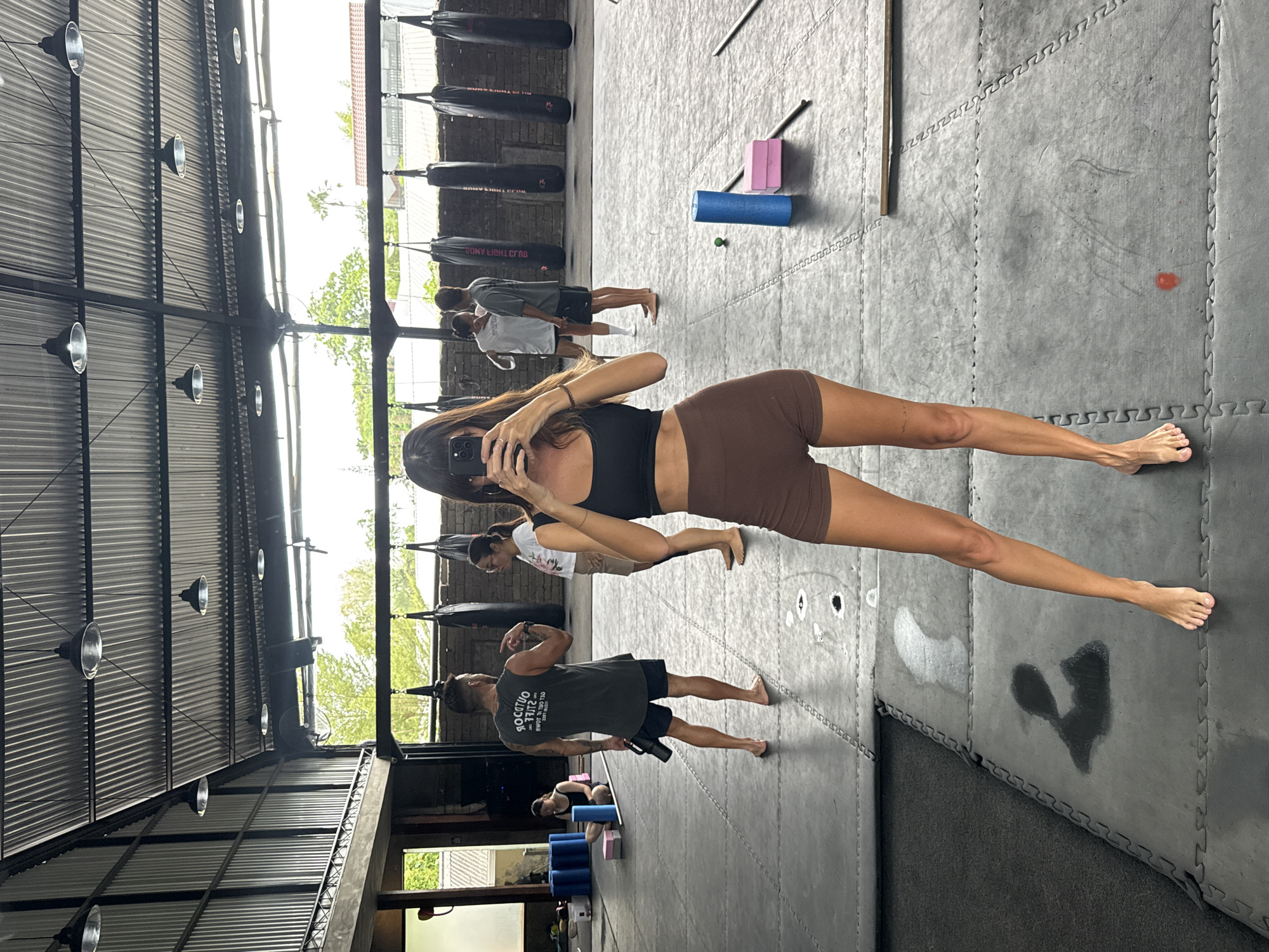 An indoor fitness studio or gym in North Kuta, Indonesia, is depicted in a brightly lit image. In the foreground, a woman with long, dark hair, wearing a black sports bra and brown athletic shorts, stands barefoot while taking a mirror selfie with her phone, which obscures part of her face.
The gym features a high corrugated metal ceiling from which industrial-style dome lights hang. Large windows line one side, allowing abundant natural light to stream in and offering a glimpse of green trees and building rooftops outside, indicating it is daytime. The floor is covered with grey interlocking foam mats.
Several other individuals are present in the gym. Two people stand close together in the mid-ground, one in a grey t-shirt and the other in a white t-shirt, both barefoot. Further back, a man in a dark sleeveless top is seen flexing an arm while holding a dark water bottle. Another person is partially visible near a stack of blue yoga blocks and mats.
Exercise equipment includes multiple black punching bags hanging against a brick wall. Other visible items are pink and blue yoga blocks, a blue foam roller, and a small green ball. Dark wet spots, suggestive of sweat, are visible on the grey floor mats, with one notable mark resembling a heart shape. Text on the punching bags appears to read "SONY LIGHT CLUB," and mirrored text on the man's sleeveless top reads "OUTDOOR LIFE."