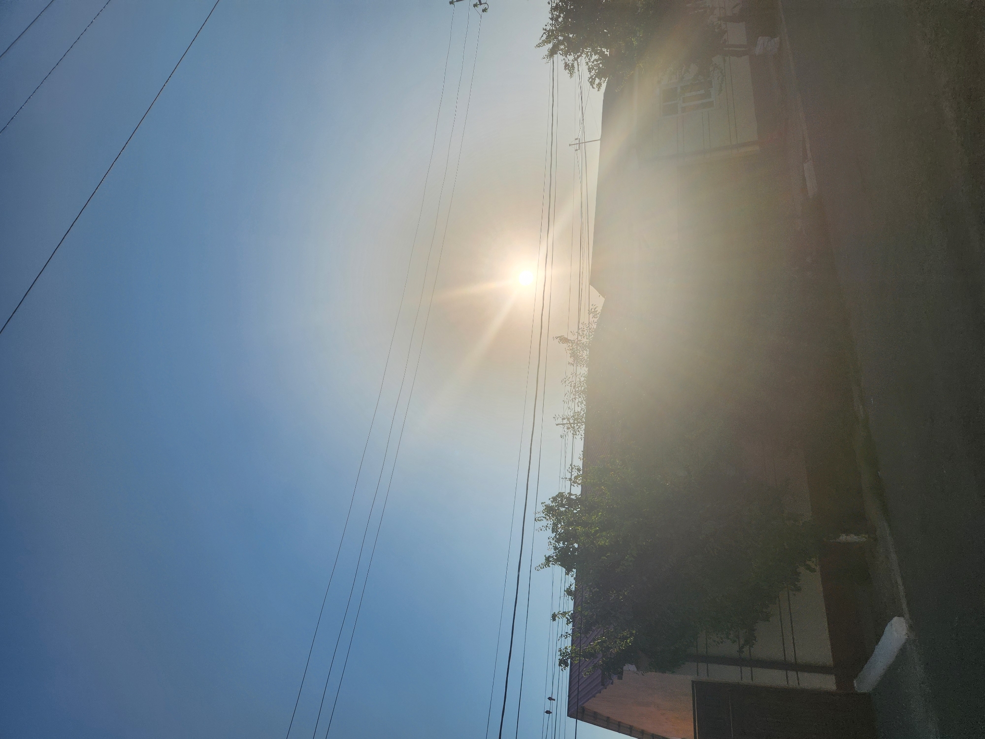 The image depicts a clear, bright sky over a residential area. The sun is visible, casting a strong glare and lens flare across the scene. Several power lines are strung across the sky. In the foreground, partially obscured by the sun's glare and shadows, are the roofs of houses, trees, and a paved road. The architecture suggests a typical suburban or rural setting. The overall lighting indicates it is daytime, likely morning or late afternoon, given the low angle of the sun. The image does not contain any discernible individuals or explicit actions.