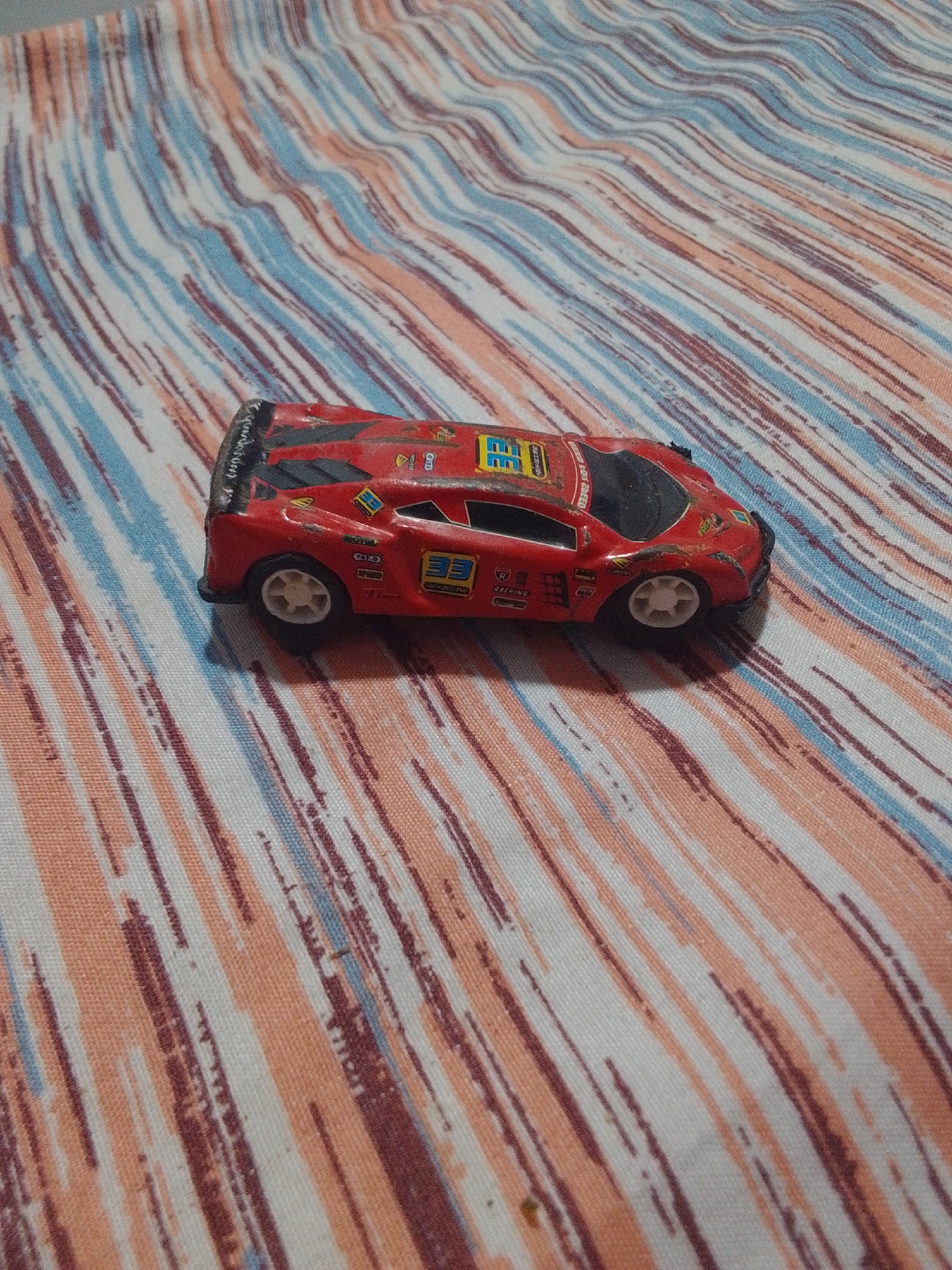 A red toy race car sits on a patterned fabric surface. The fabric has a diagonal stripe pattern in shades of white, pink, blue, and dark red. The car is positioned on its side, with the front of the car facing towards the right side of the image. The car appears to be made of plastic and has decals on its sides, including numbers and sponsor logos. The lighting suggests an indoor setting, possibly a home environment. There is no indication of the city of La Tagua, Venezuela, within the image itself.