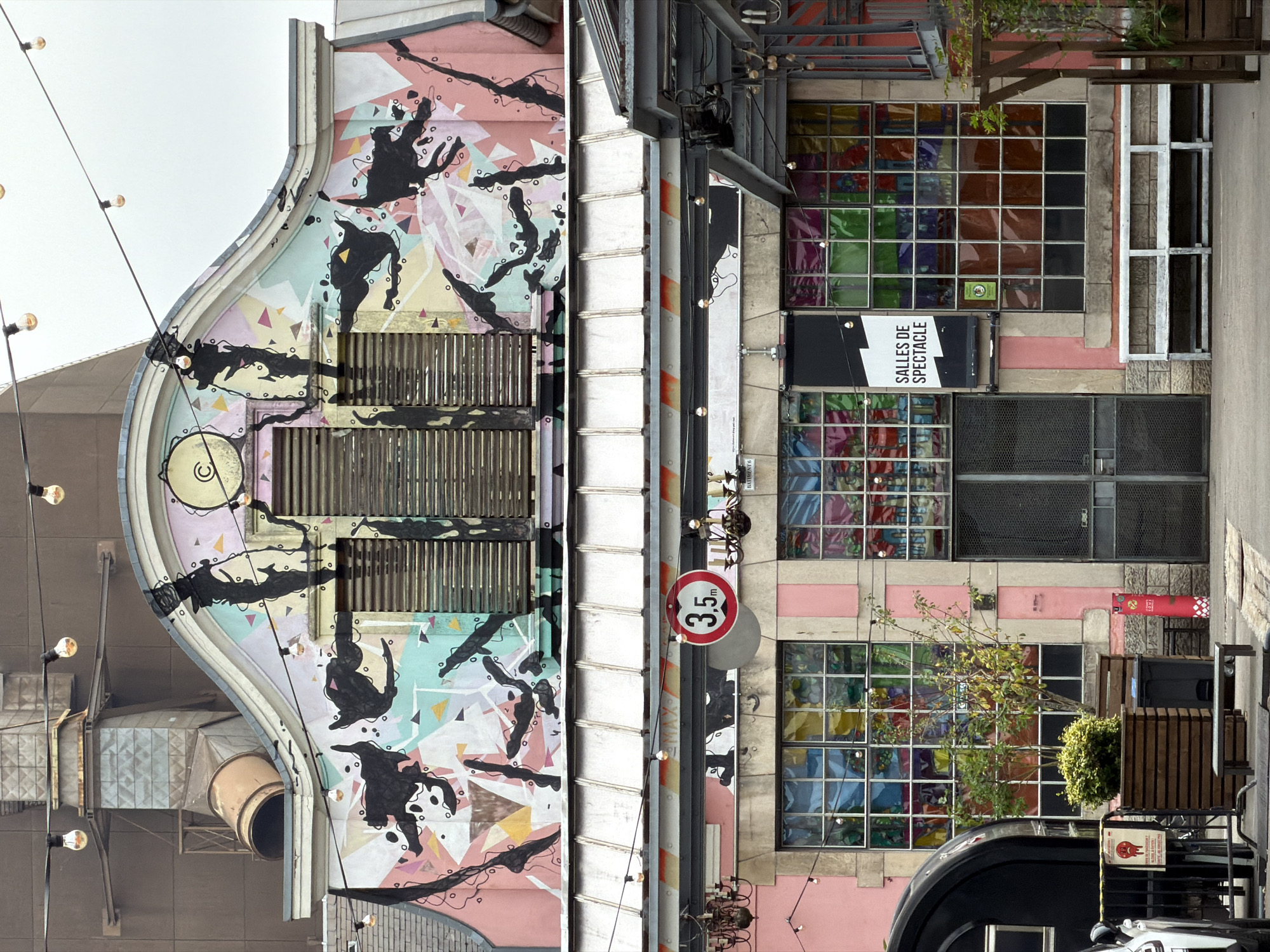 The image shows the exterior of a building with a pink facade decorated with colorful abstract art and black painted figures. The upper part of the building features a curved roofline with a large circular element at the apex. Several strings of bare light bulbs are strung across the facade. Below the art, there are two levels of windows. The upper level has louvered shutters. The lower level features large windows with multicolored stained glass. A sign reading "SALLES DE SPECTACLE" is mounted on the facade. In front of the building, there is a paved area with planters, benches, and some outdoor seating. A portion of a black vehicle is visible on the left side of the image. The overall impression is of a vibrant, arts-focused venue. The building is identified as the Rockhal, a music venue located in Esch-sur-Alzette, Luxembourg.