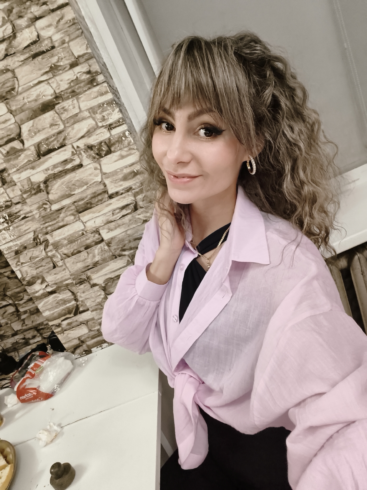 The image is a selfie of a woman with curly brown hair and bangs. She is wearing a pink button-down shirt tied at the waist over a black top. She has gold hoop earrings and a gold necklace. Her left hand is raised to her neck. Behind her is a textured wall resembling stacked stones. To her right is a window frame. On a white surface below, there is a package of napkins, food in a bowl, and a chocolate shaped like a heart. Based on online data, the location is Chaikovsky, Russia.