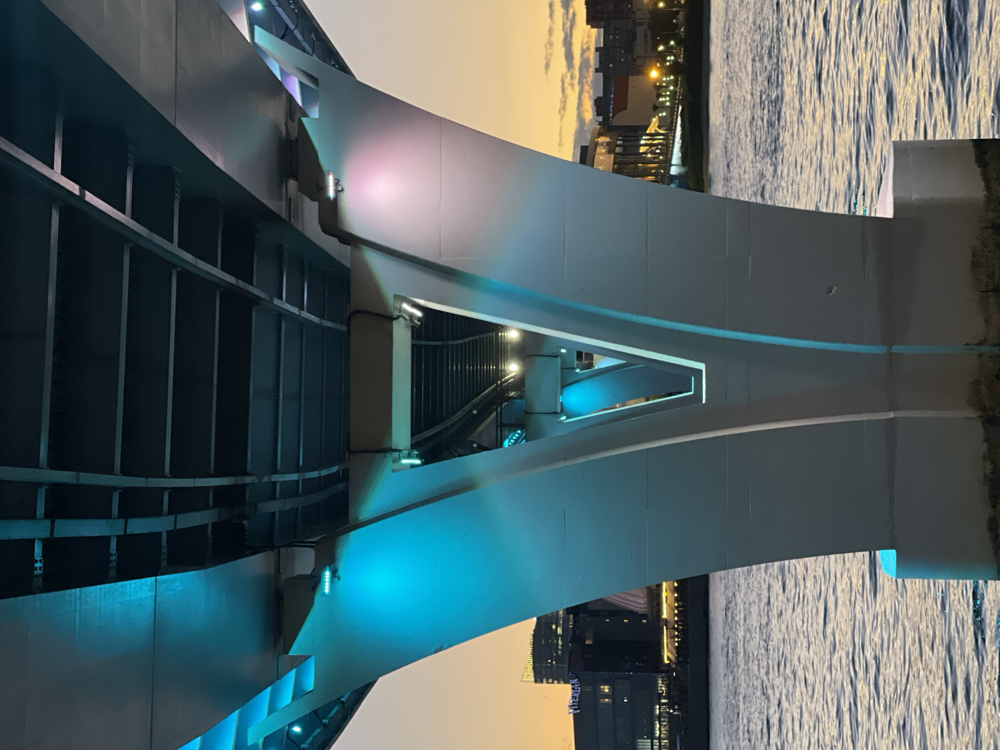 An upward perspective shot of the underside of a modern bridge illuminated with blue and pink lights, at sunset. The bridge structure is composed of large, white concrete supports that curve upwards, meeting the dark metal framework of the bridge's deck. The water below reflects the lights and the warm hues of the sunset. In the background, city buildings and lights are visible on the horizon, with the sky displaying a gradient of orange and yellow. No people are present in the frame.