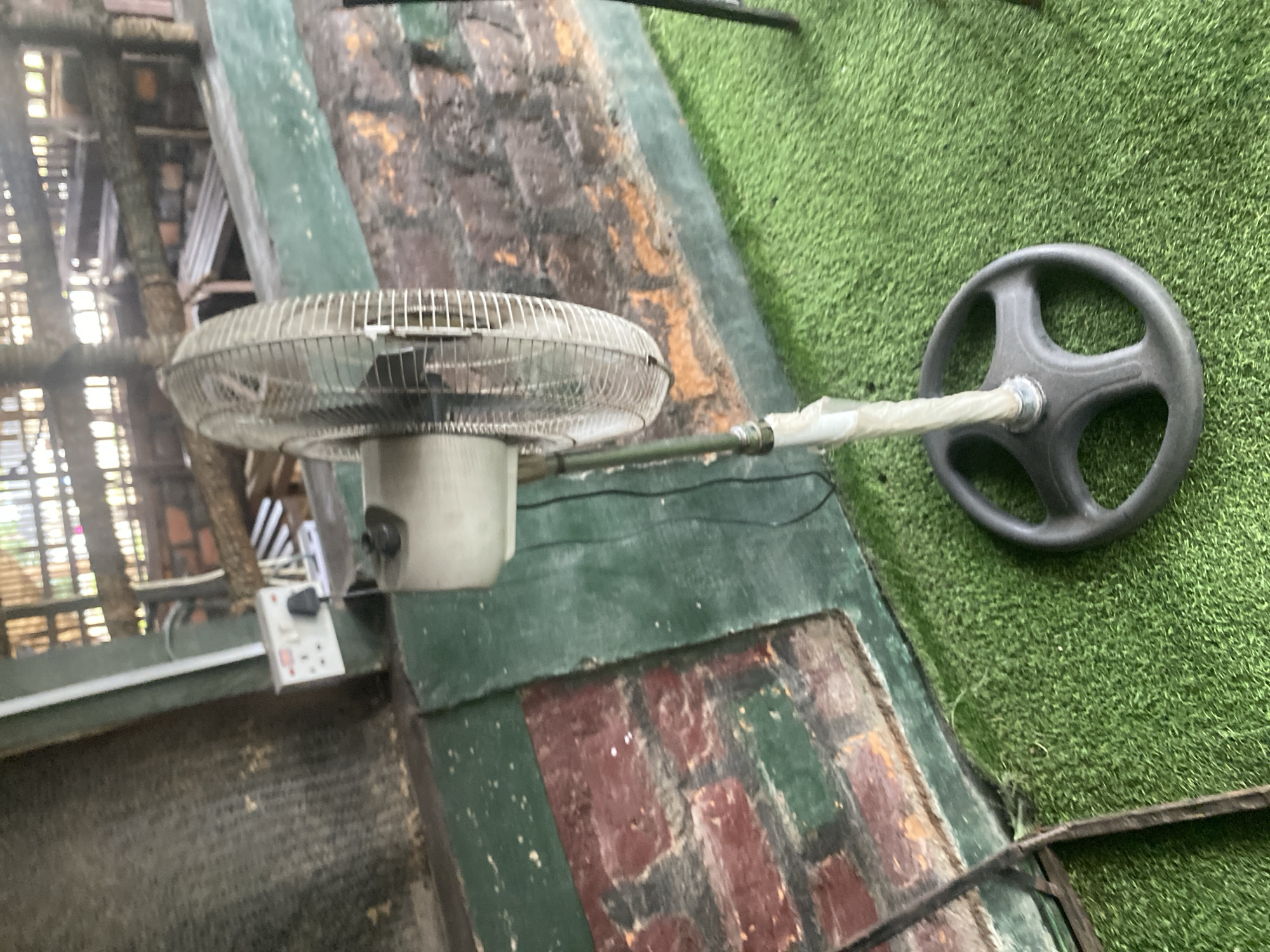 An outdoor scene is depicted with a standing fan on a green artificial turf. The fan is silver and white, with a round base and a metal grill. In the background, there is a brick wall painted green and red, with wooden structures above. A white electrical outlet with plugs is visible on the wall. The overall setting appears to be an outdoor area, possibly a backyard or a seating area in Jalingo, Nigeria. There are no people or animals present in the image. The lighting suggests it is daytime.