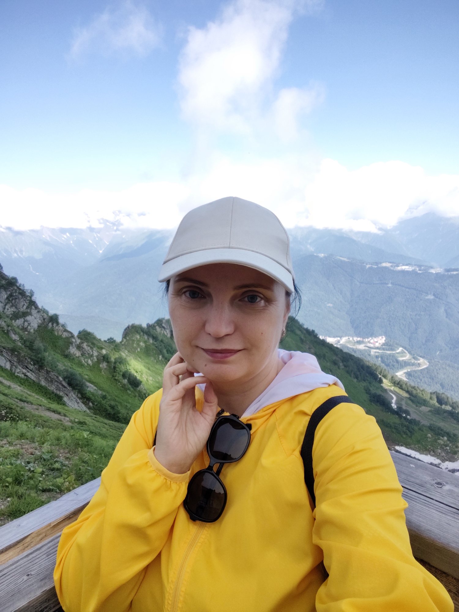 A woman wearing a white baseball cap and a bright yellow hooded jacket poses for a selfie in a mountainous landscape. She is standing on a wooden platform, with lush green slopes and distant snow-capped mountains visible in the background under a bright blue sky with scattered clouds. A winding road is visible in the valley below, and a small village is nestled on the mountainside. The woman has dark hair and is looking at the camera with a slight smile, her hand touching her chin. She has sunglasses hanging from the zipper of her jacket. The image appears to have been taken during the daytime, with clear weather suggesting a pleasant day for outdoor activities. The location is identified as Estosadok, Russia, a known mountain resort area.